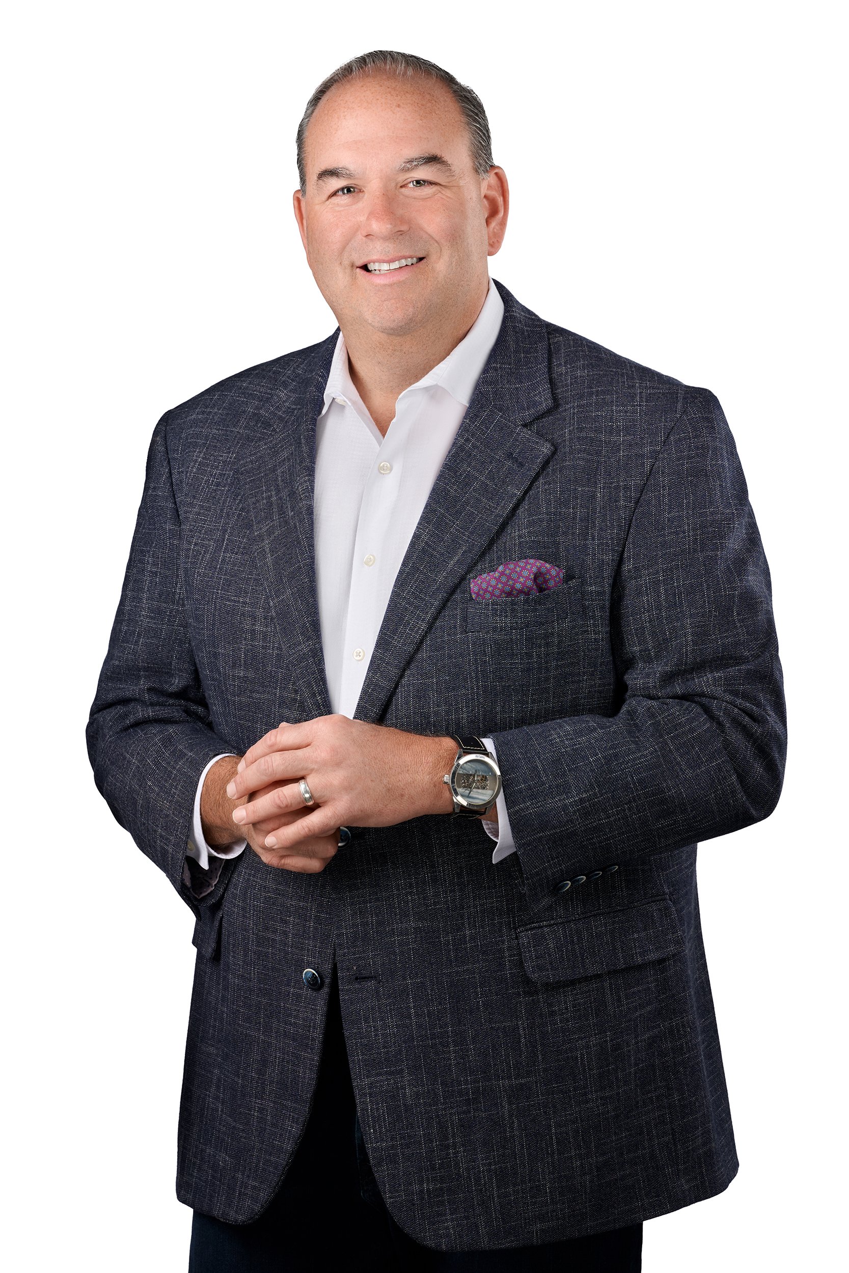 A man in a dark blazer with a white shirt, smiling, standing against a white background.