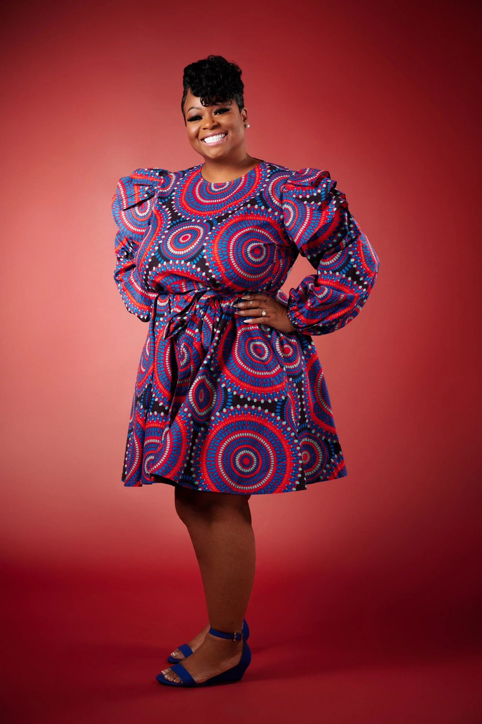 A smiling woman with short curly hair wearing a red, blue, and black patterned dress with puffed sleeves, standing against a red background.