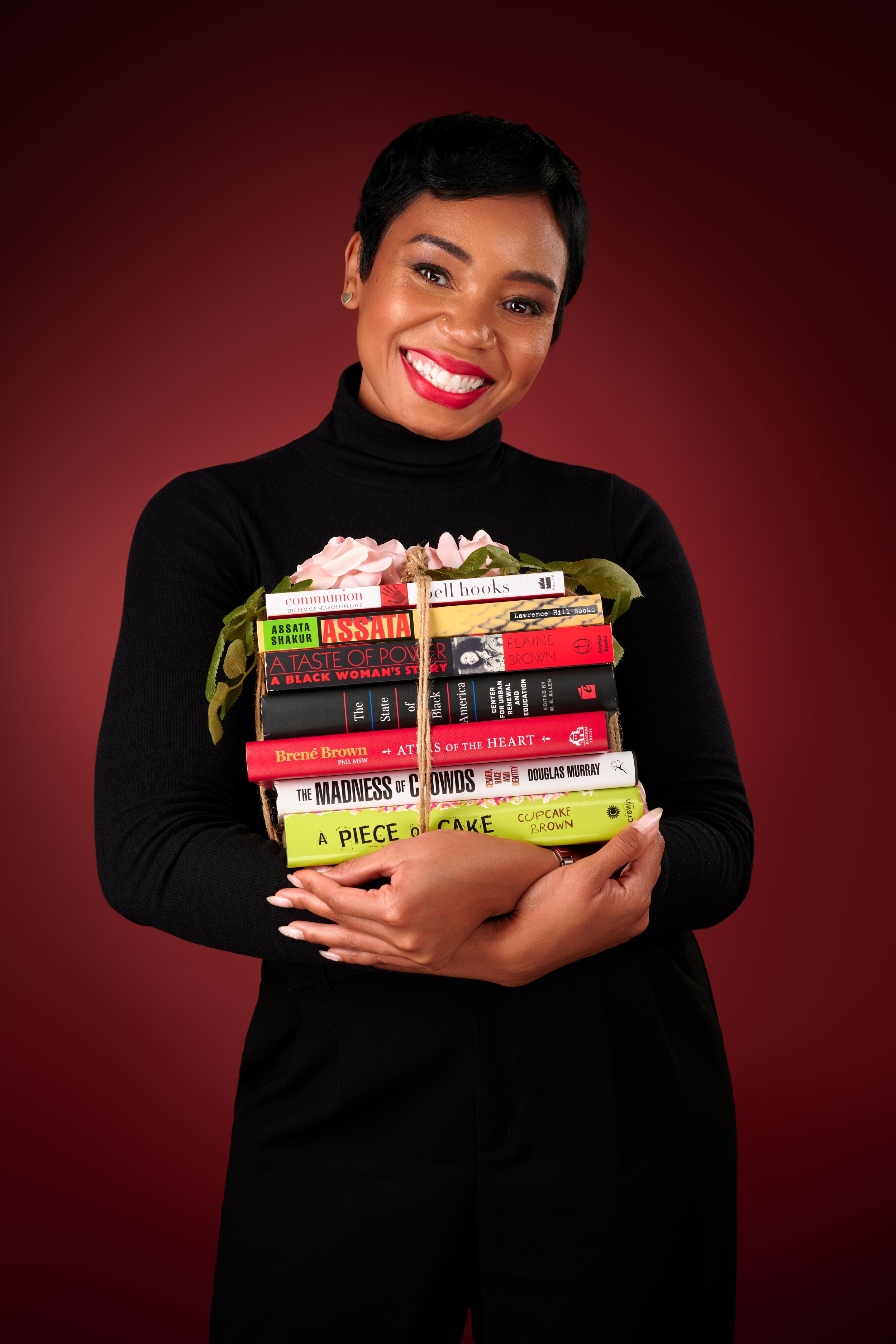 A woman wearing a black turtleneck, smiling, holding a bundle of books with pink flowers on top against a dark red background.