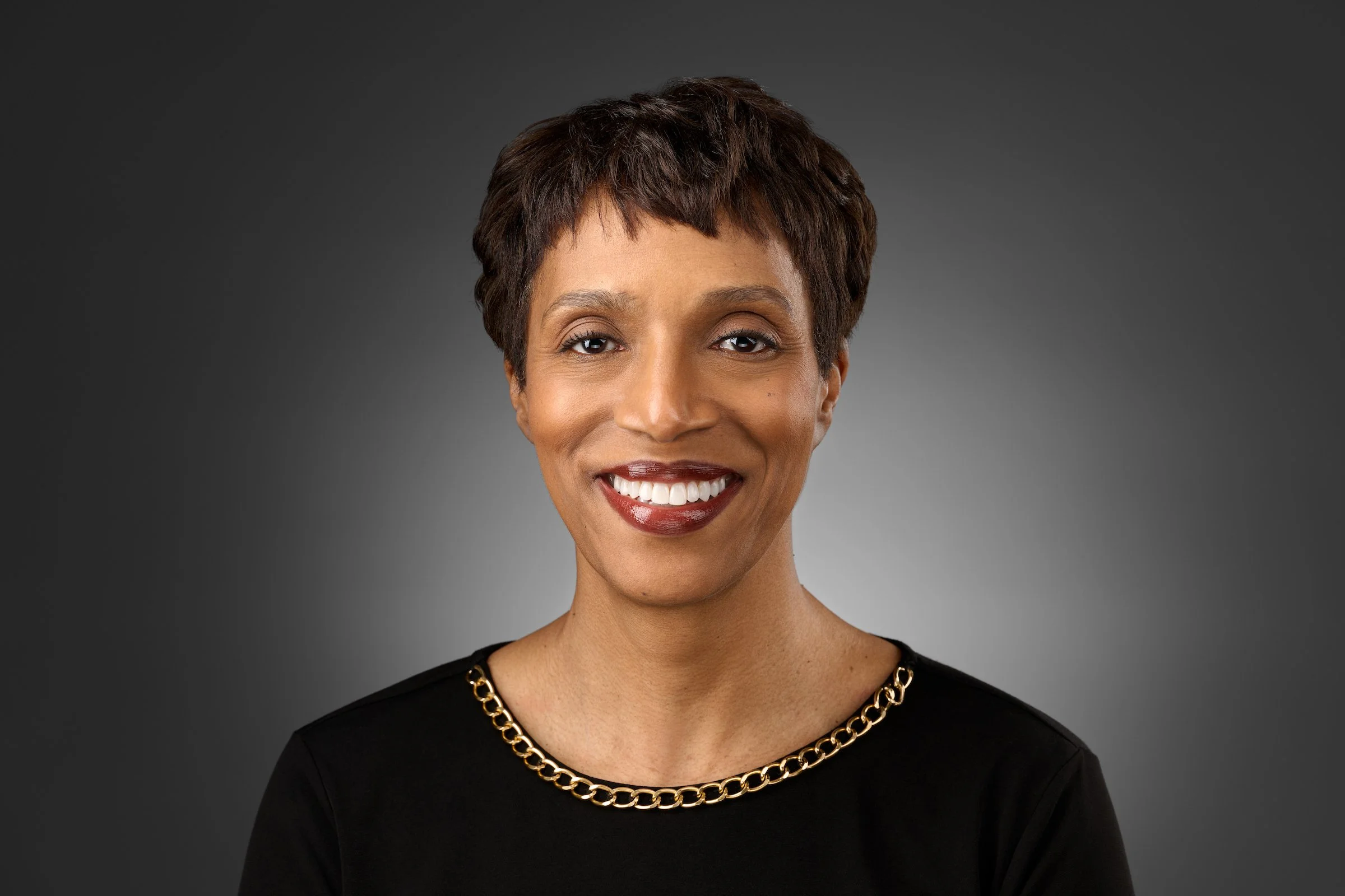 A woman with short, dark hair wearing a black top with a gold chain neckline, smiling at the camera against a dark gray background.