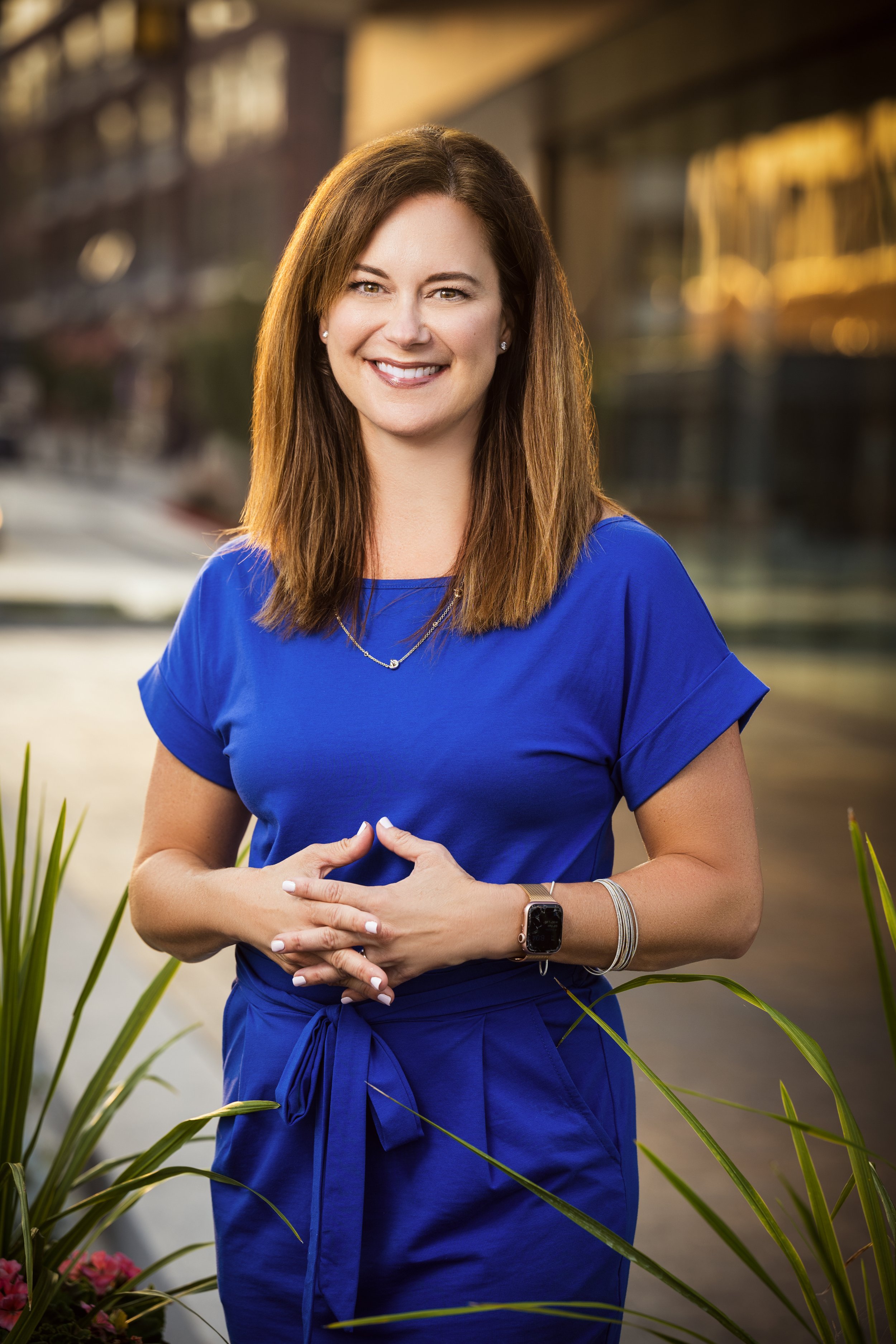 A smiling woman with brown hair, wearing a blue dress, standing outdoors near modern buildings, with plants in the foreground.
