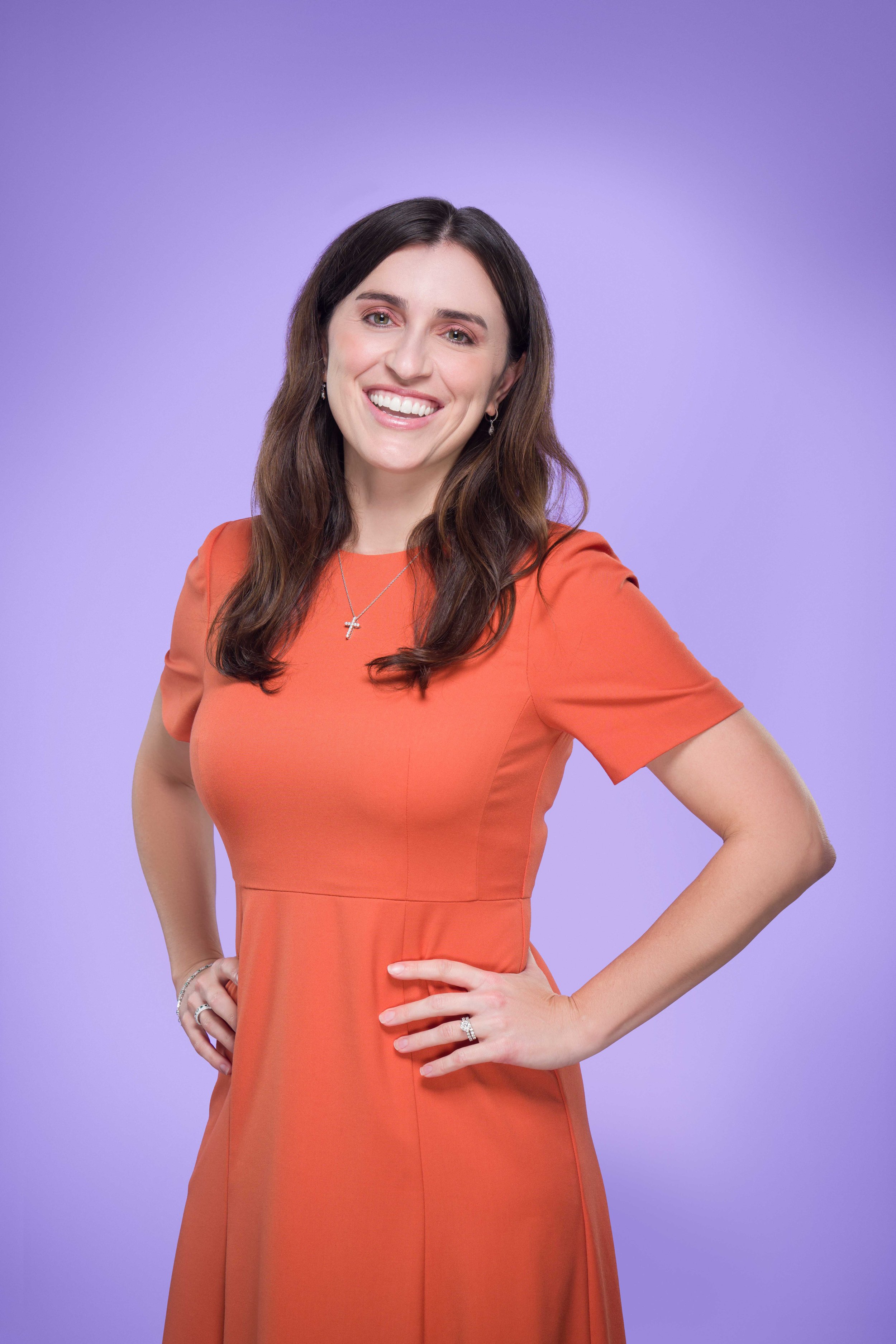 A woman with long brown hair wearing an orange dress, smiling with her hands on her hips, standing against a purple background.