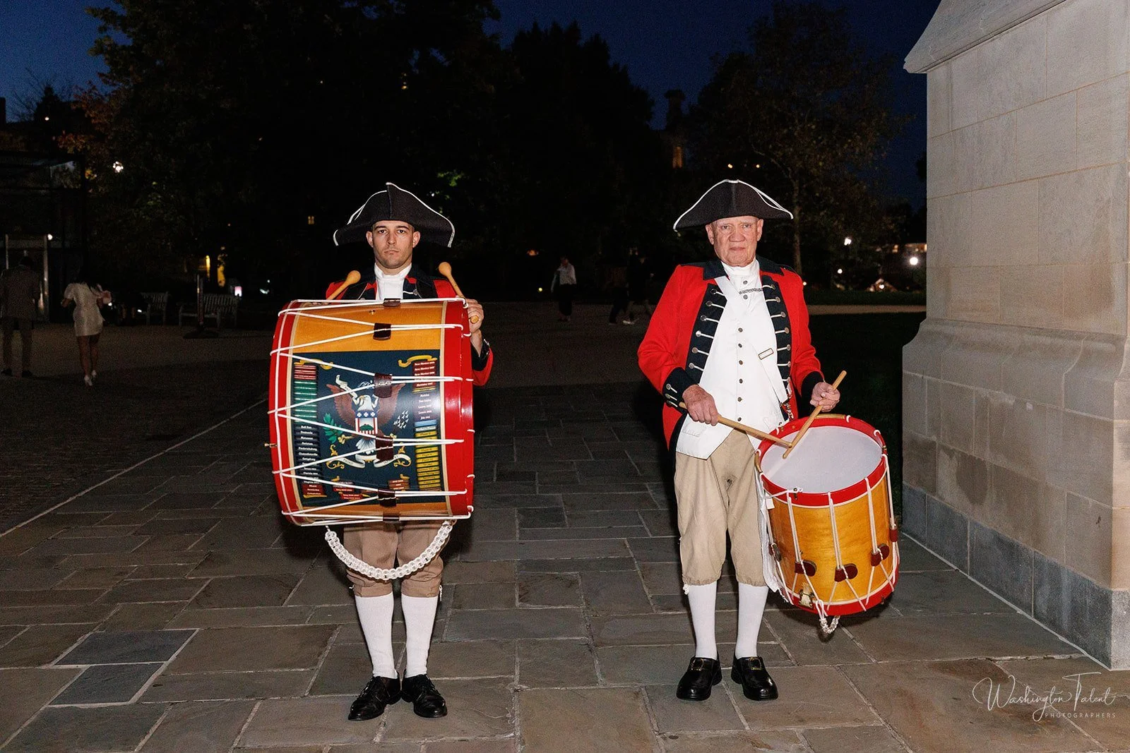 Two men dressed as Revolutionary War soldiers, each holding a drum, standing on a paved area at night.
