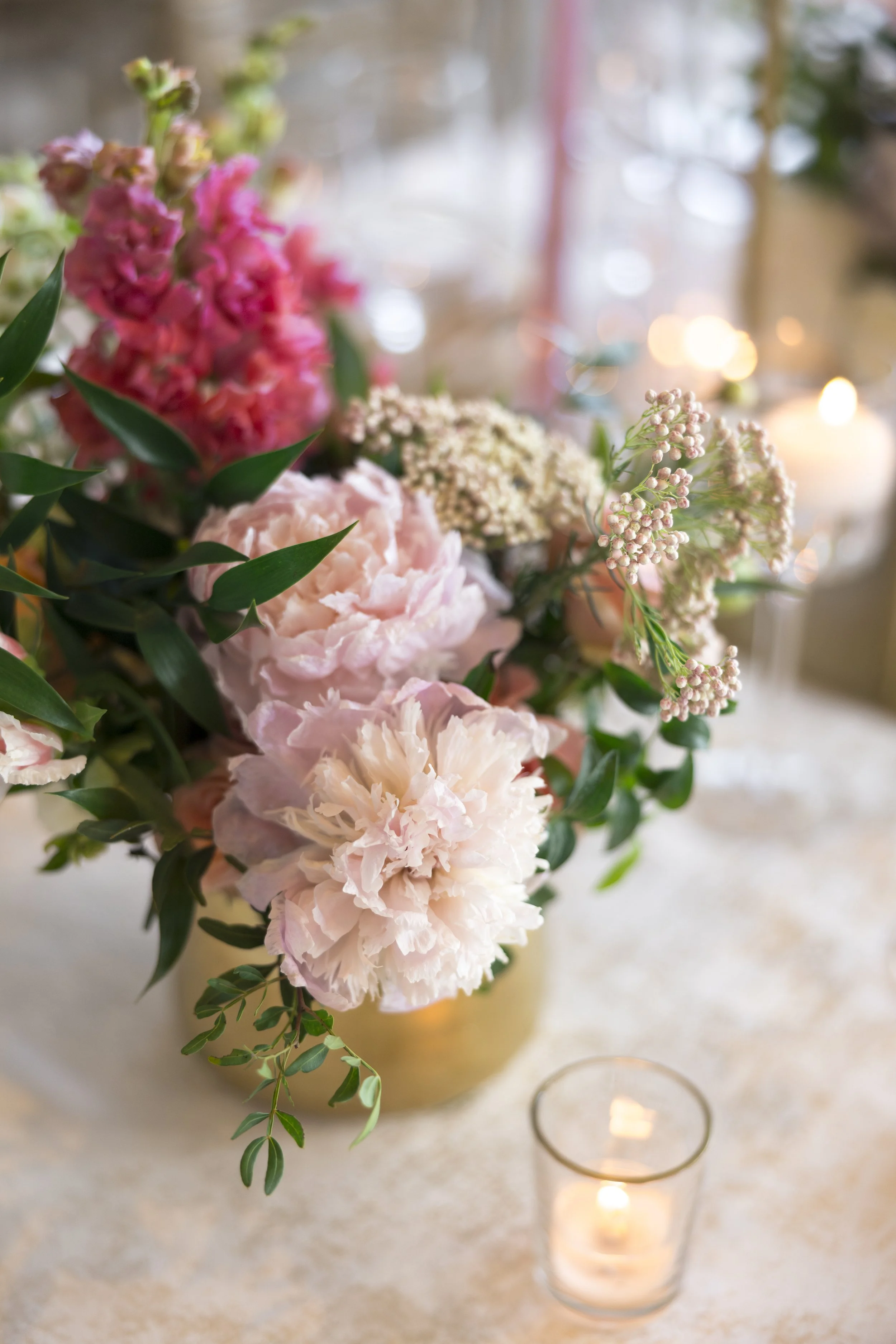 Pink and white peonies and other flowers in a gold vase with teal leaves, with a lit candle in a glass holder on a marble surface.