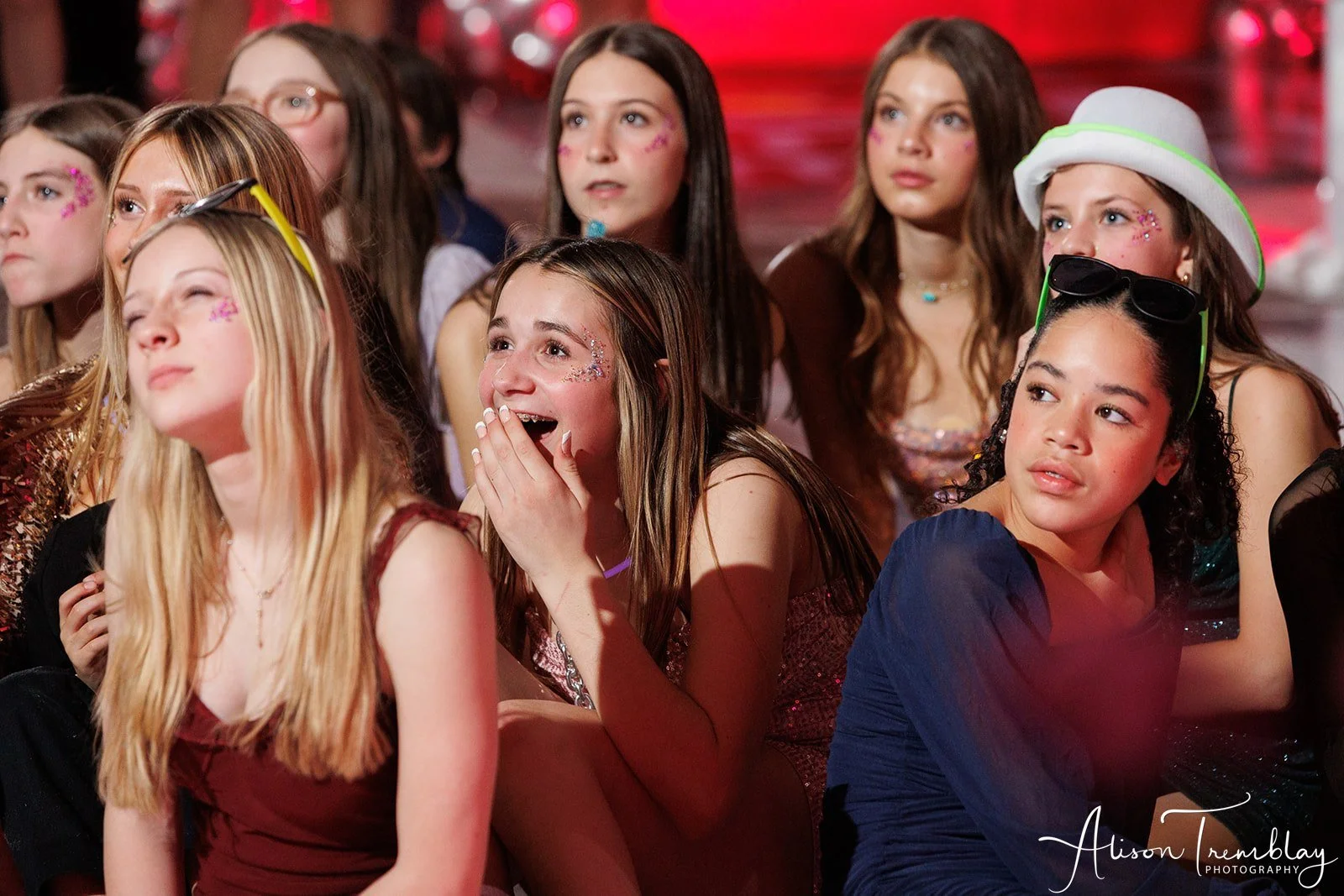 Group of young girls at a party, some smiling and some surprised, with colorful face decorations and party accessories like glasses and hats.