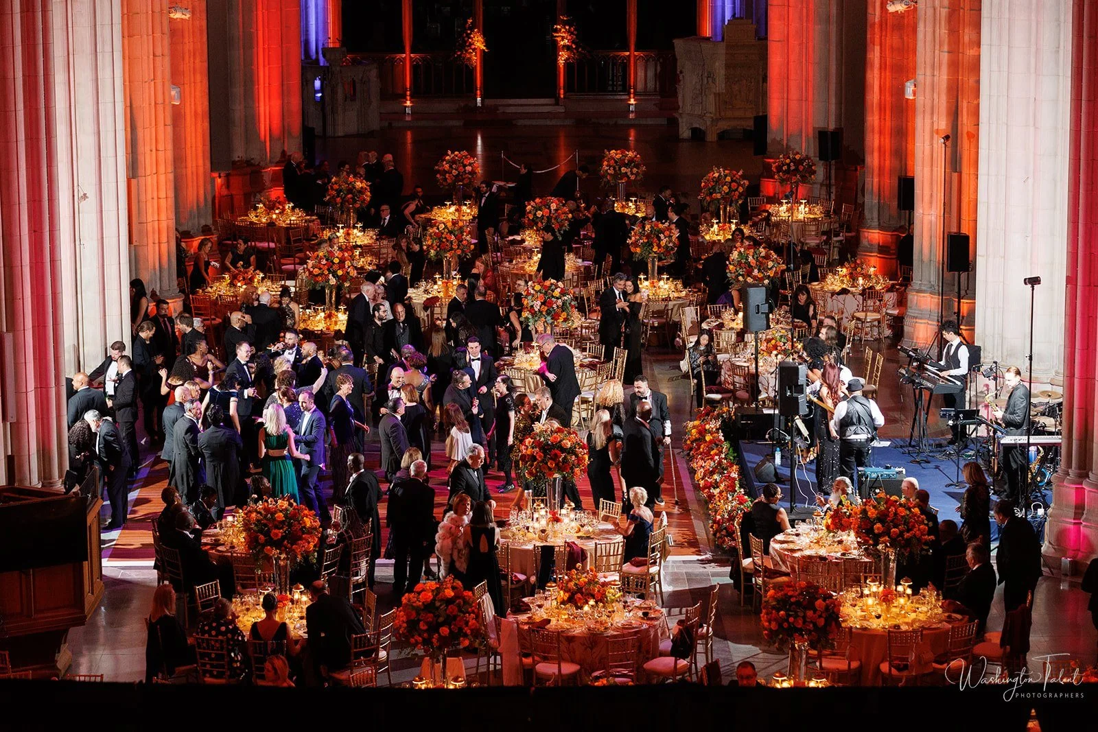 Guests dancing and socializing at a formal event with decorated tables and a live band in a grand hall with tall columns and colorful lighting.