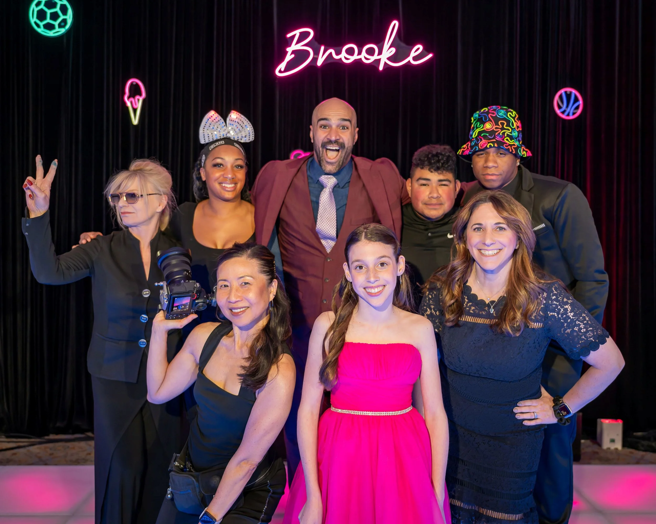 Group of people posing for a photo at a party with a black curtain backdrop and neon signs, including a pink 'Brooke' sign, ice cream, basketball, and soccer ball graphics.
