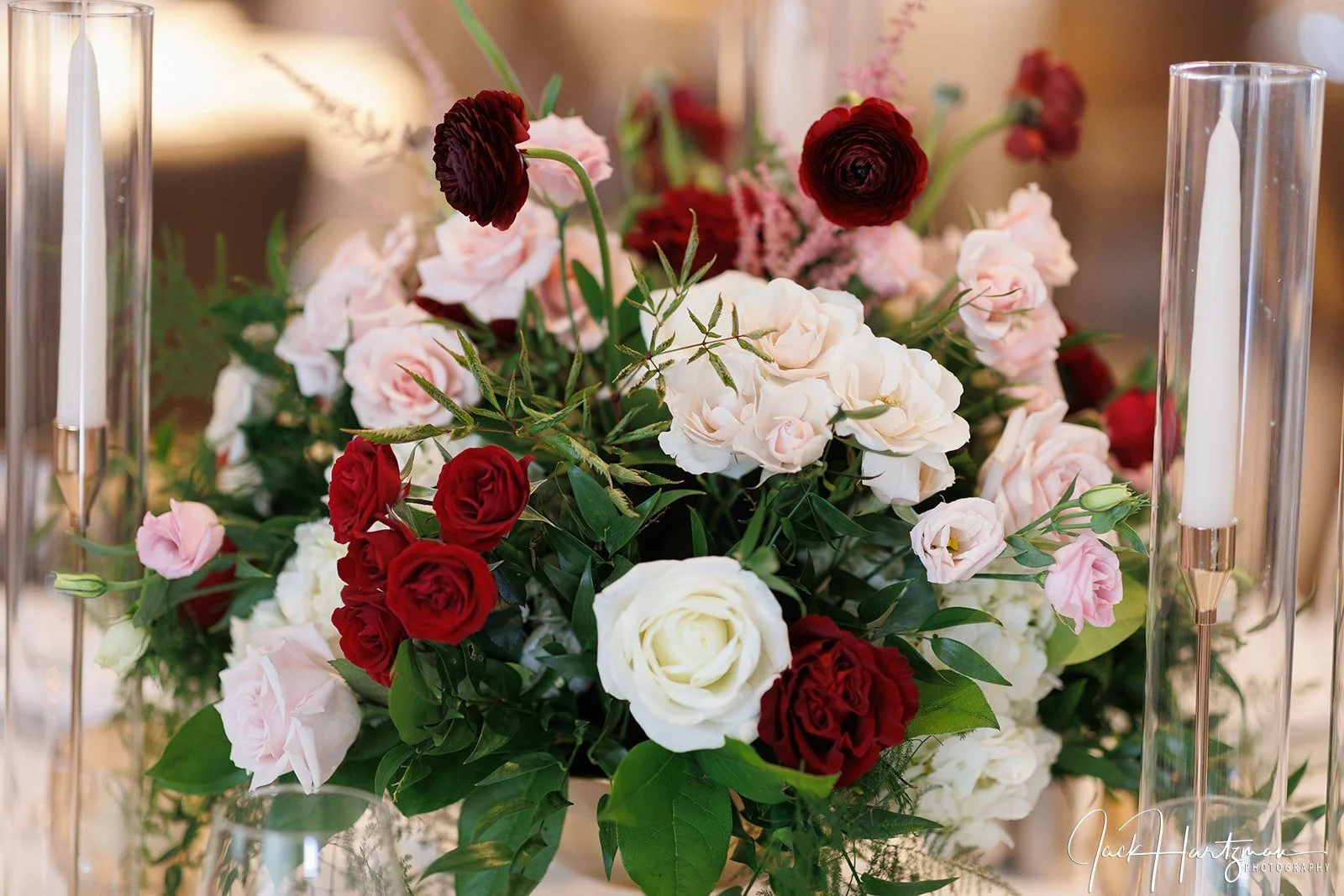 A floral arrangement with pink, red, and white roses and dark red ranunculus flowers, surrounded by green leaves, flanked by tall glass candle holders with white candles.