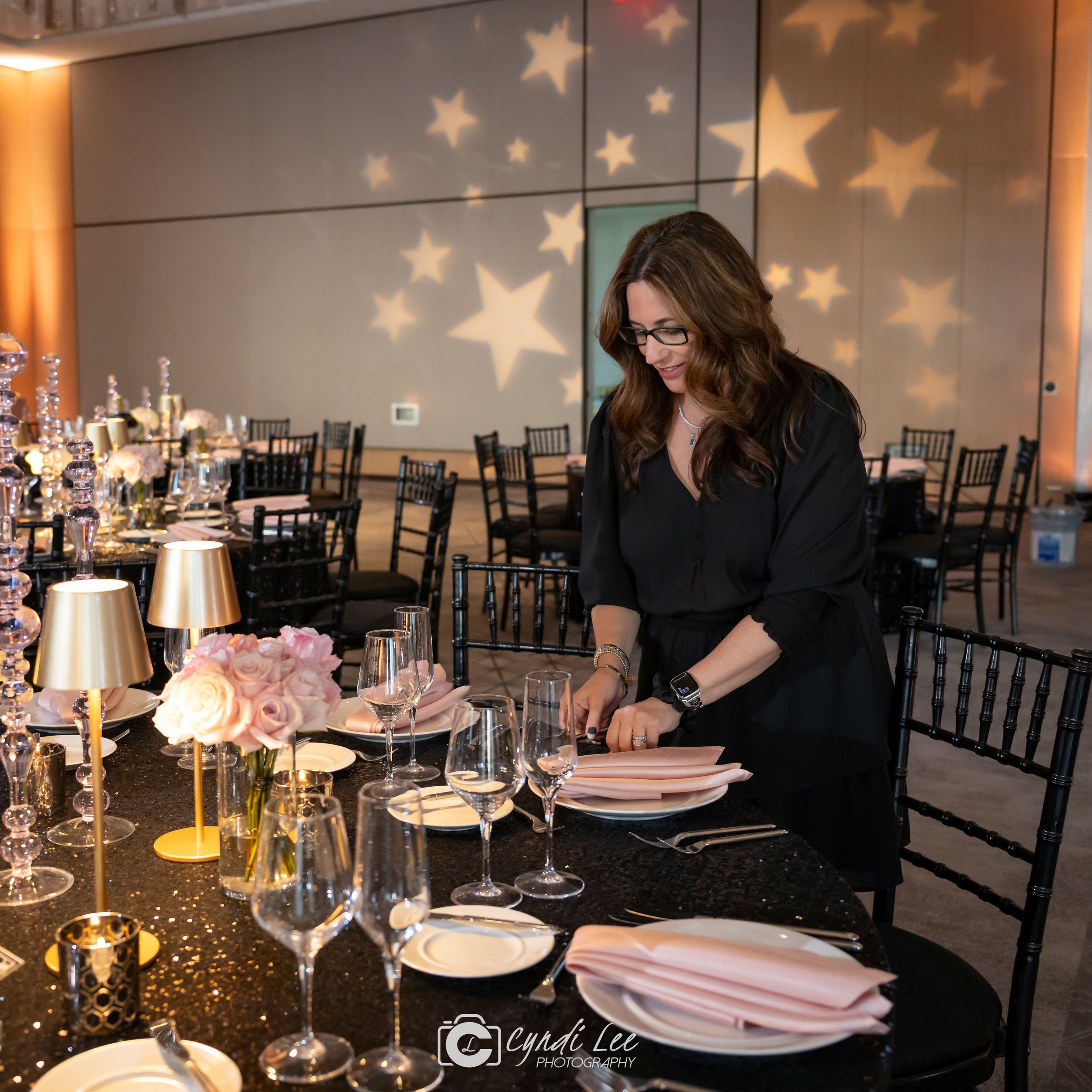 Woman in black dress preparing table with pink napkins in a decorated event hall with star-patterned lighting on the walls.