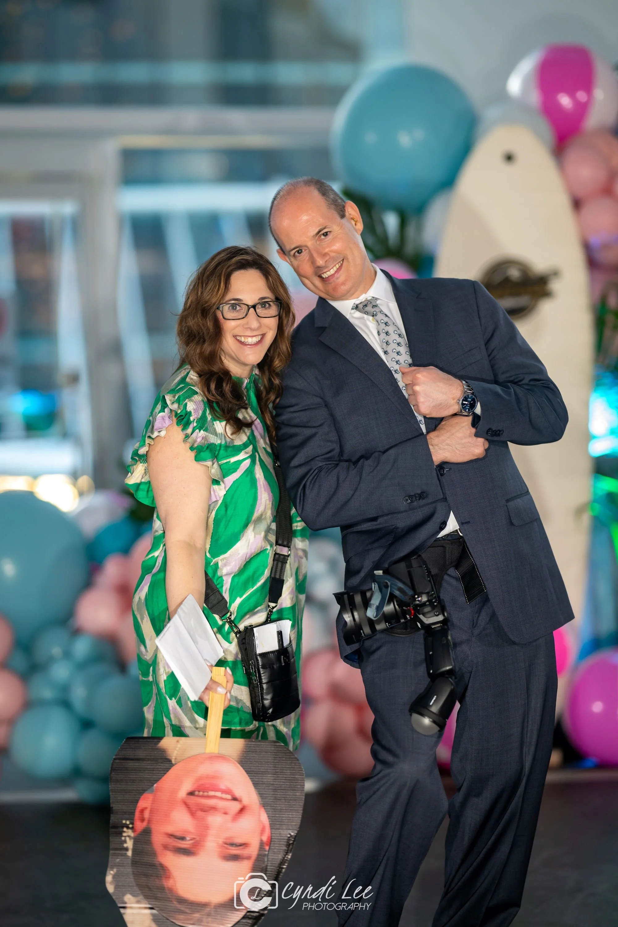 A woman and a man standing together indoors during a celebration, with colorful balloons in the background. The woman is wearing glasses and a green patterned dress, holding a paddle with her upside-down photo on it. The man is dressed in a suit, smiling, with a camera hanging from his belt.