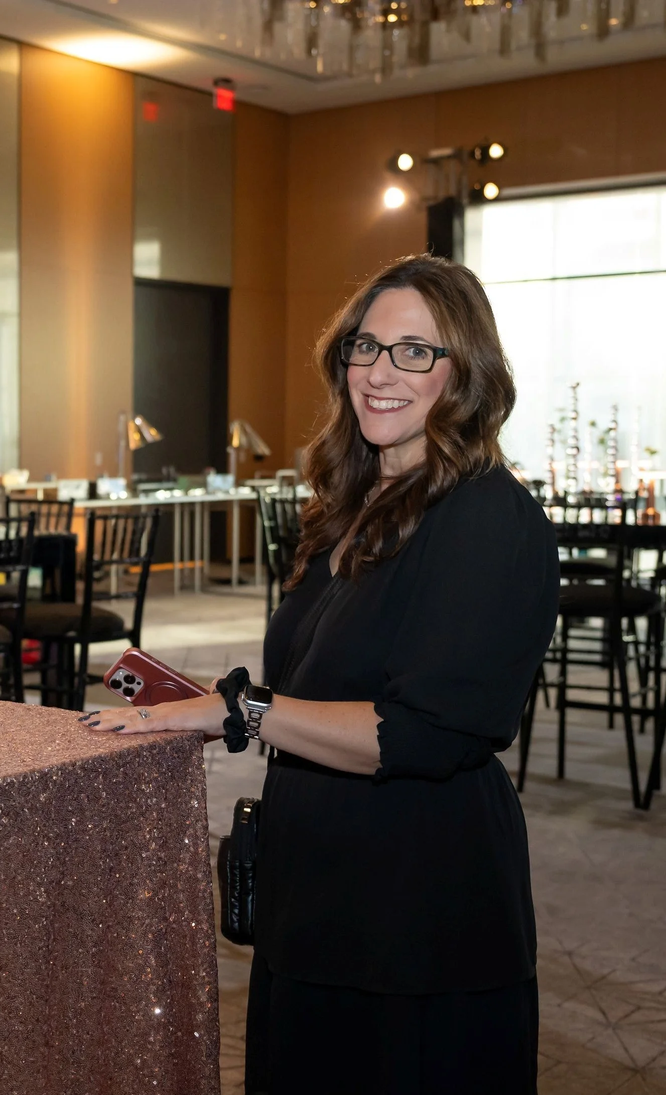 A woman with brown wavy hair, glasses, and a black dress standing at a decorated reception desk holding a smartphone, in an event hall with tables, chairs, and chandeliers.