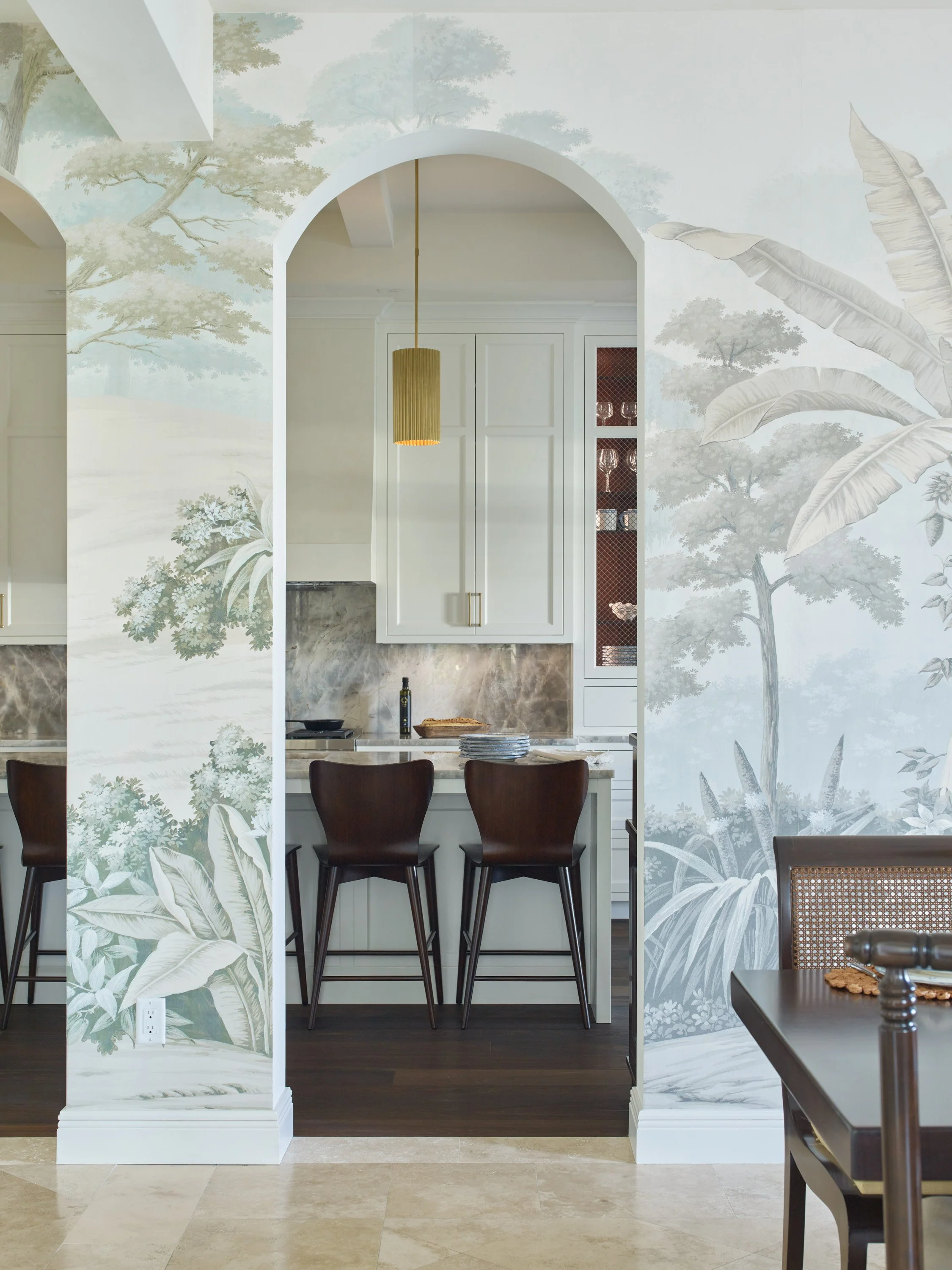 View through an archway into a kitchen with a grey marble backsplash, white cabinets, dark wood stools at a countertop, and gold pendant lights.