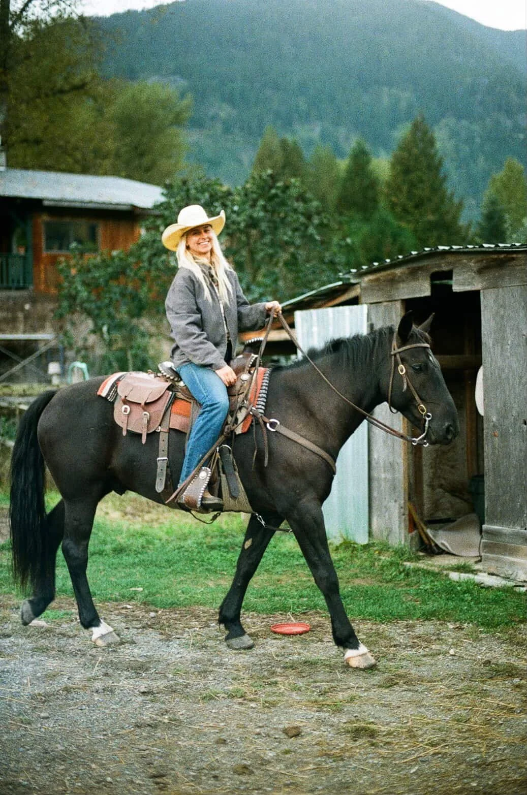 A woman with long blonde hair wearing a cowboy hat, a gray jacket, and blue jeans, riding a black horse near a wooden barn with a mountain and trees in the background.
