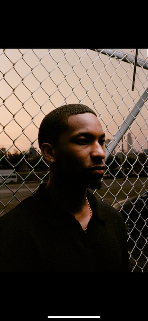 A young man with dark skin and short hair standing in front of a chain-link fence during sunset.