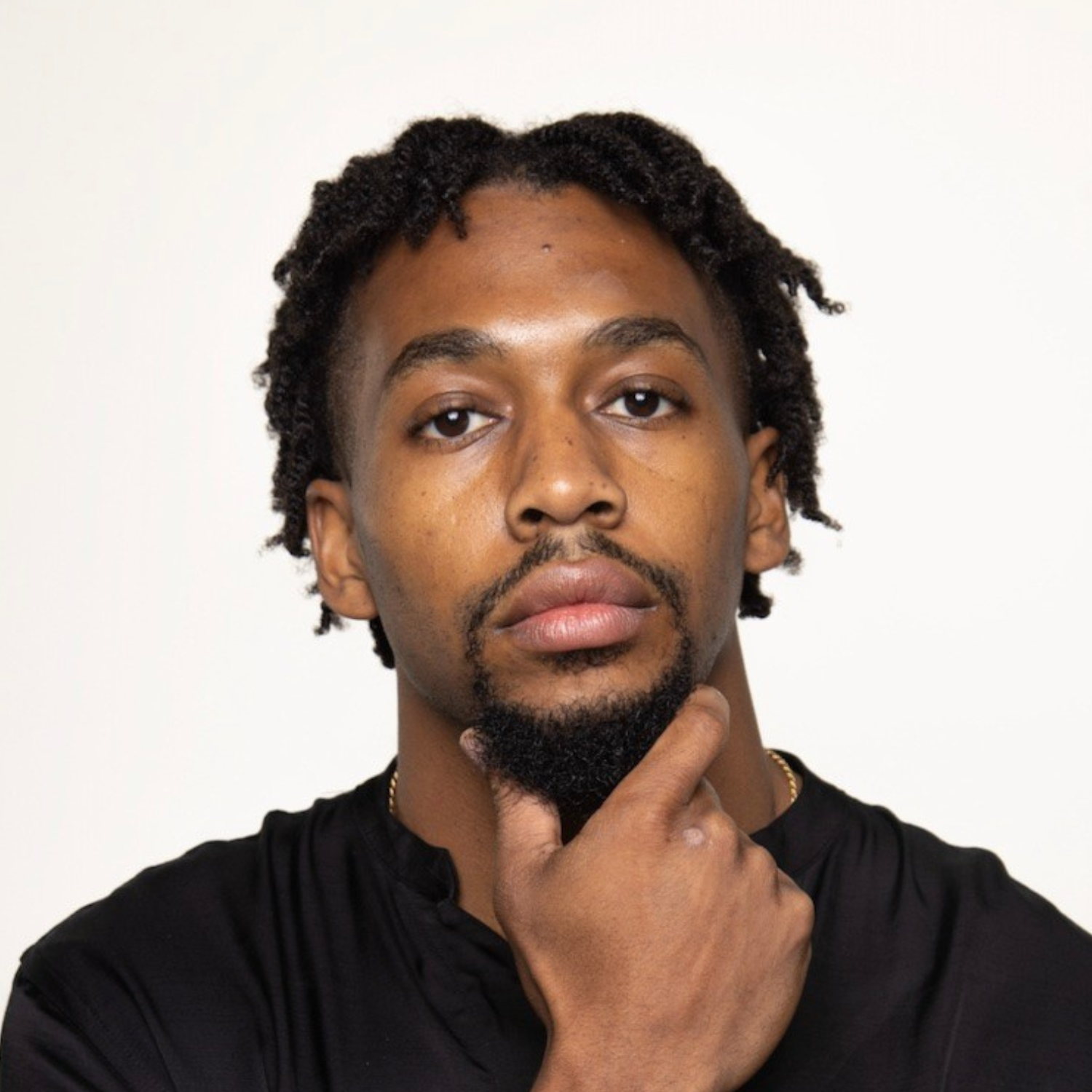 Portrait of a young Black man with short curly hair, facial hair, wearing a black shirt, holding his chin thoughtfully against a plain white background.