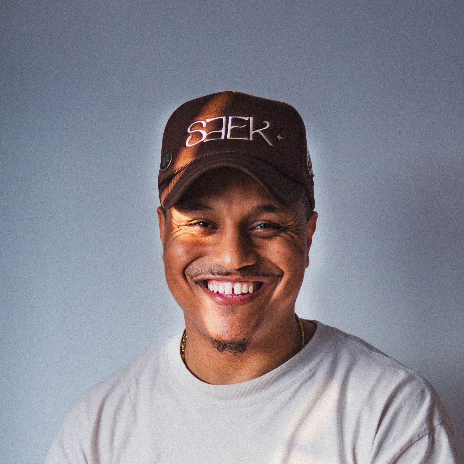 Portrait of a young Black man with short curly hair, facial hair, wearing a black shirt, holding his chin thoughtfully against a plain white background.