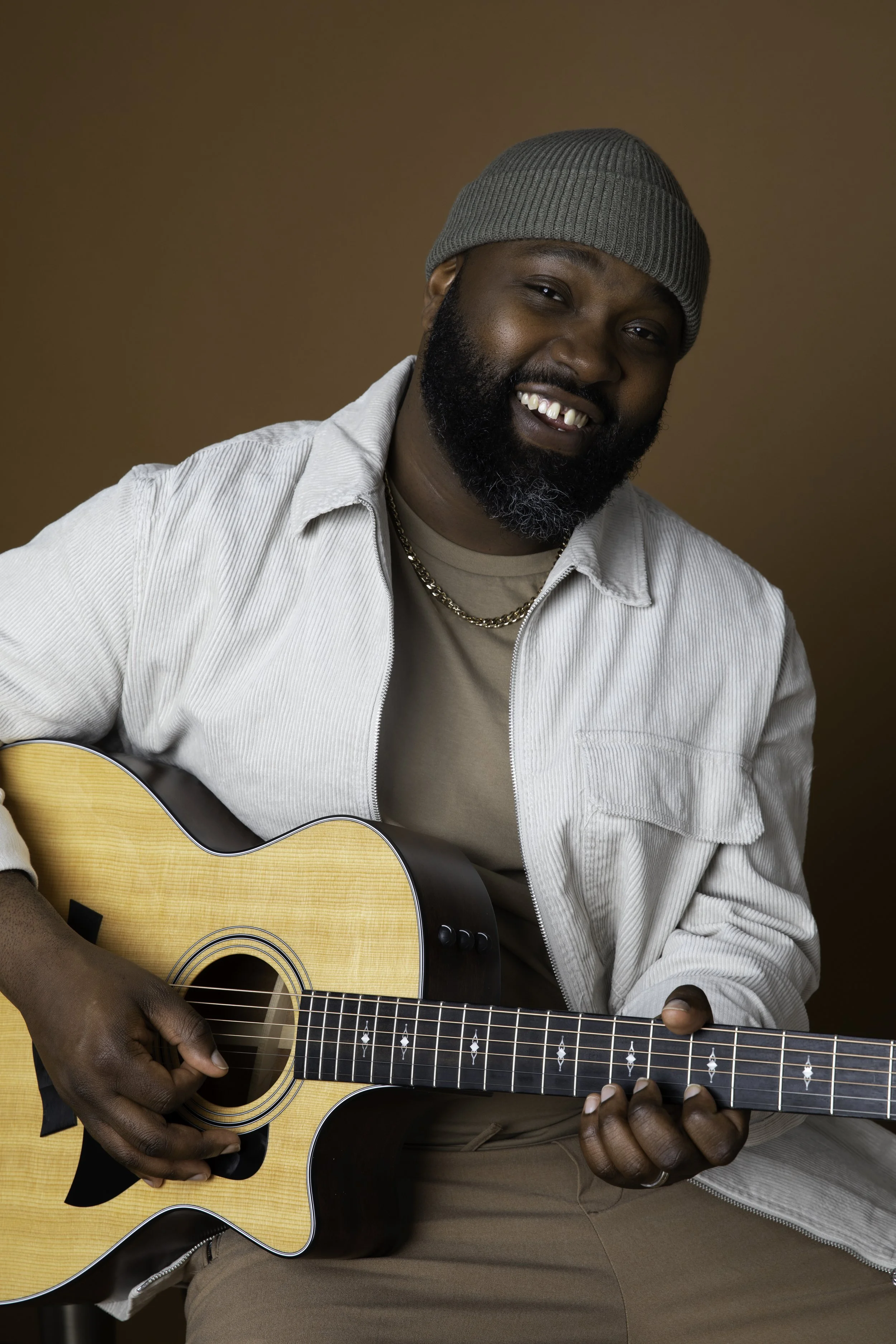 A man with a dark beard and mustache wearing a gray beanie, beige t-shirt, and white corduroy jacket playing an acoustic guitar against a brown background.