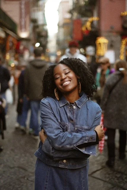 A smiling young woman with curly hair wearing a denim jacket and earrings, standing on a busy street with blurred pedestrians and buildings in the background.