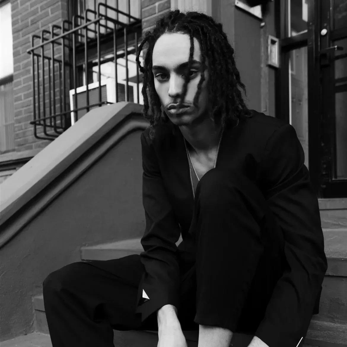 A young man with shoulder-length dreadlocks sitting on outdoor stairs, wearing dark clothing, looking directly at the camera with a serious expression, in black and white.