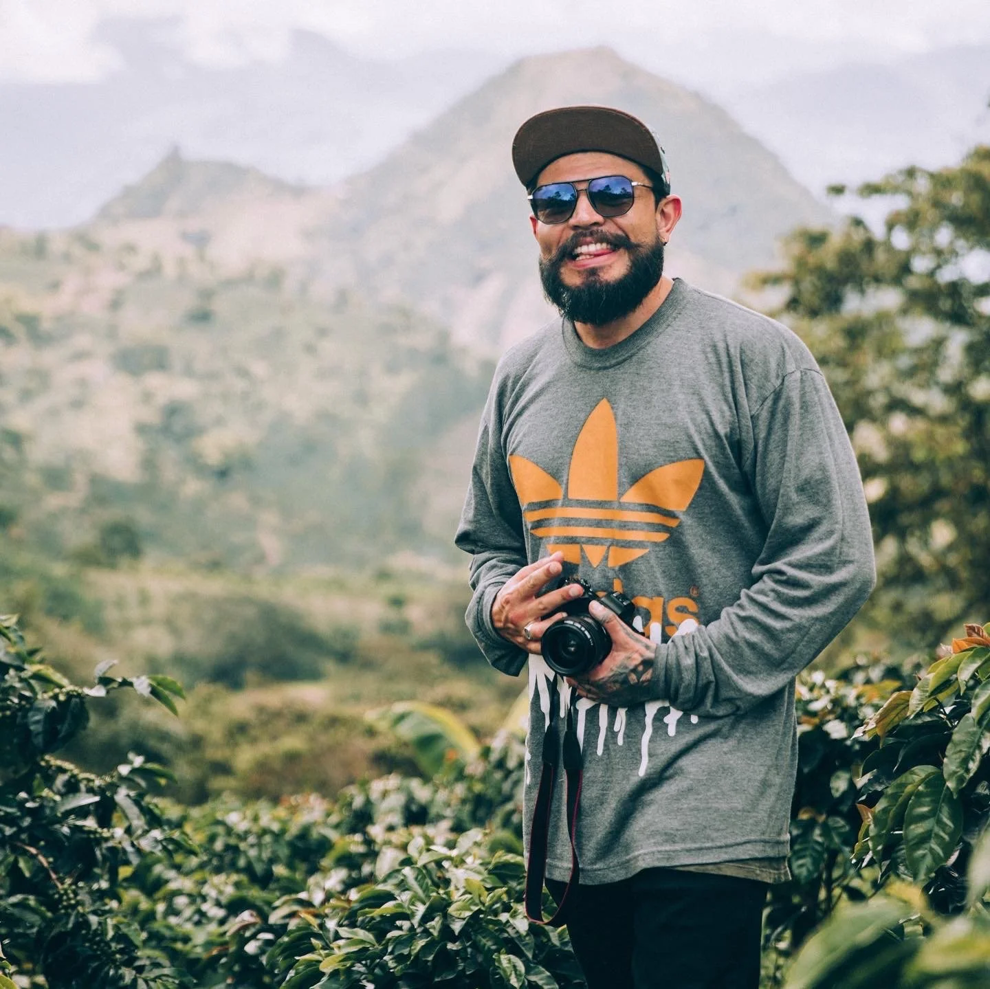 Portrait of Know Where Coffee Head Roaster and Sourcing Leader, Steven Martinez. Steven is standing in front of coffee plants in the foreground. In the background, a scenic landscape at coffee origin