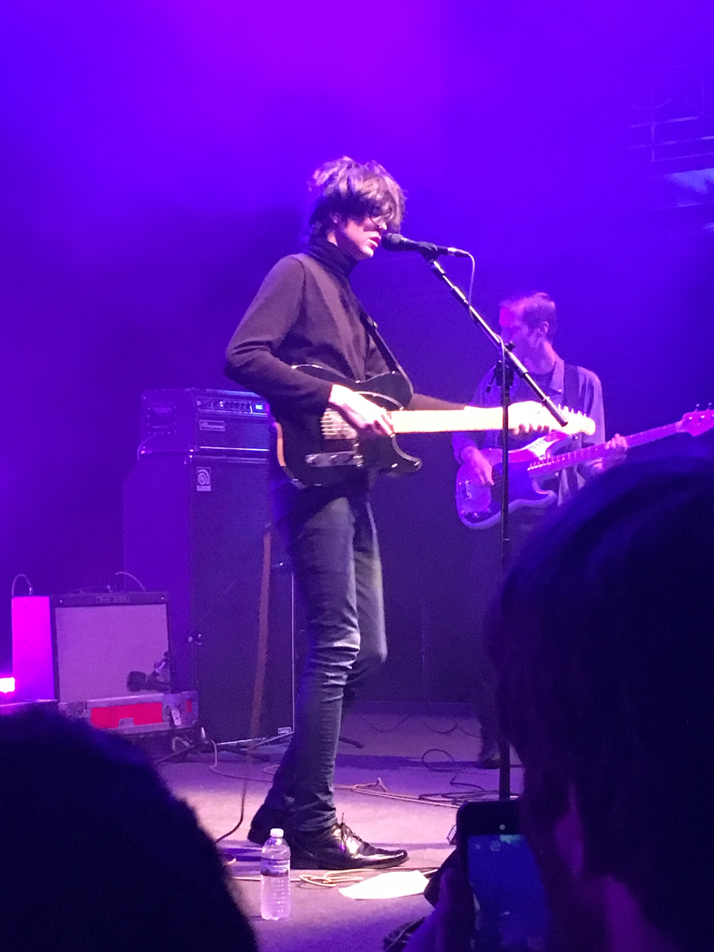 Car Seat Headrest’s Will Toledo performs at the 9:30 Club in Washington, D.C. His hair and the purple lighting cover his face. He stands at the mic and holds an electric guitar.