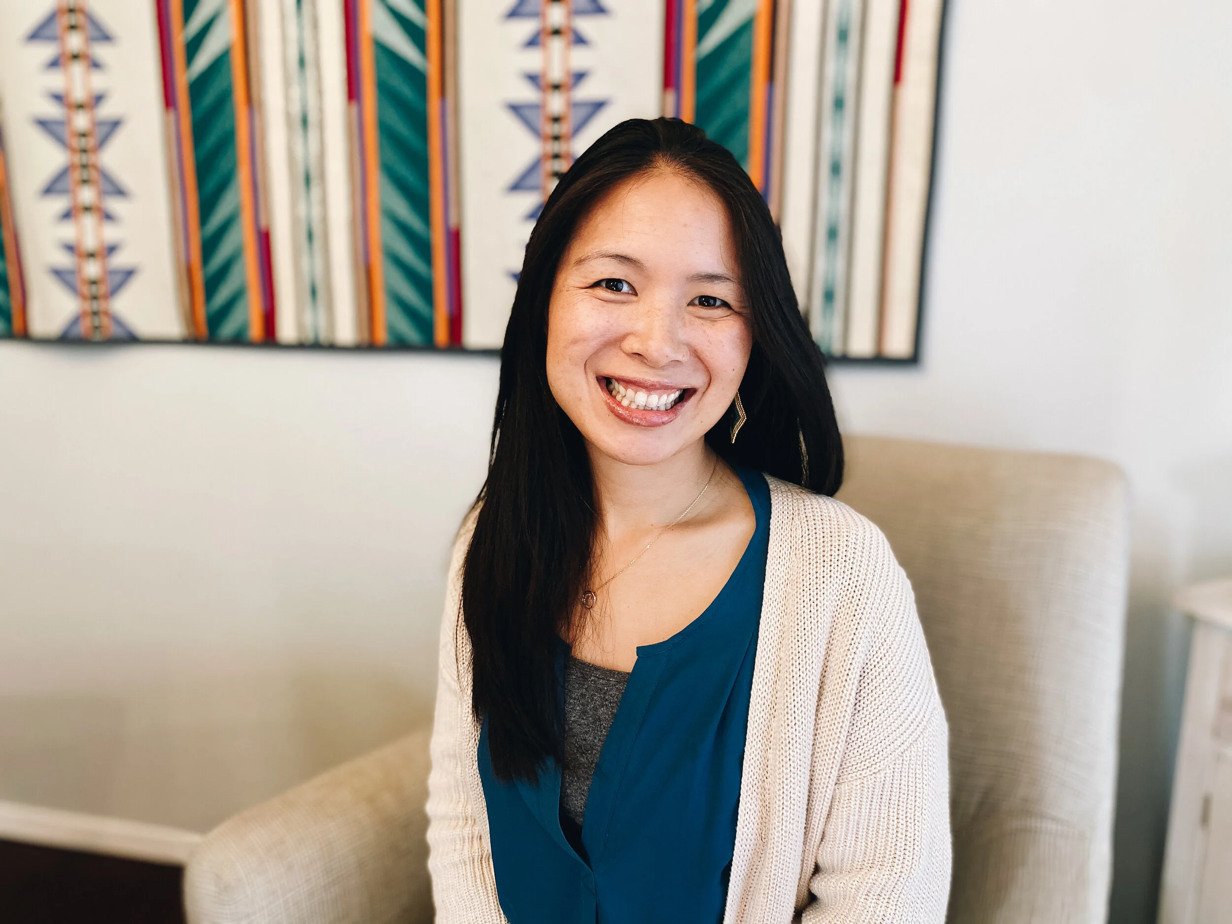 A smiling woman with long black hair, wearing a blue top and a cream-colored cardigan, sitting on a beige armchair in a room with a colorful abstract wall hanging in the background.
