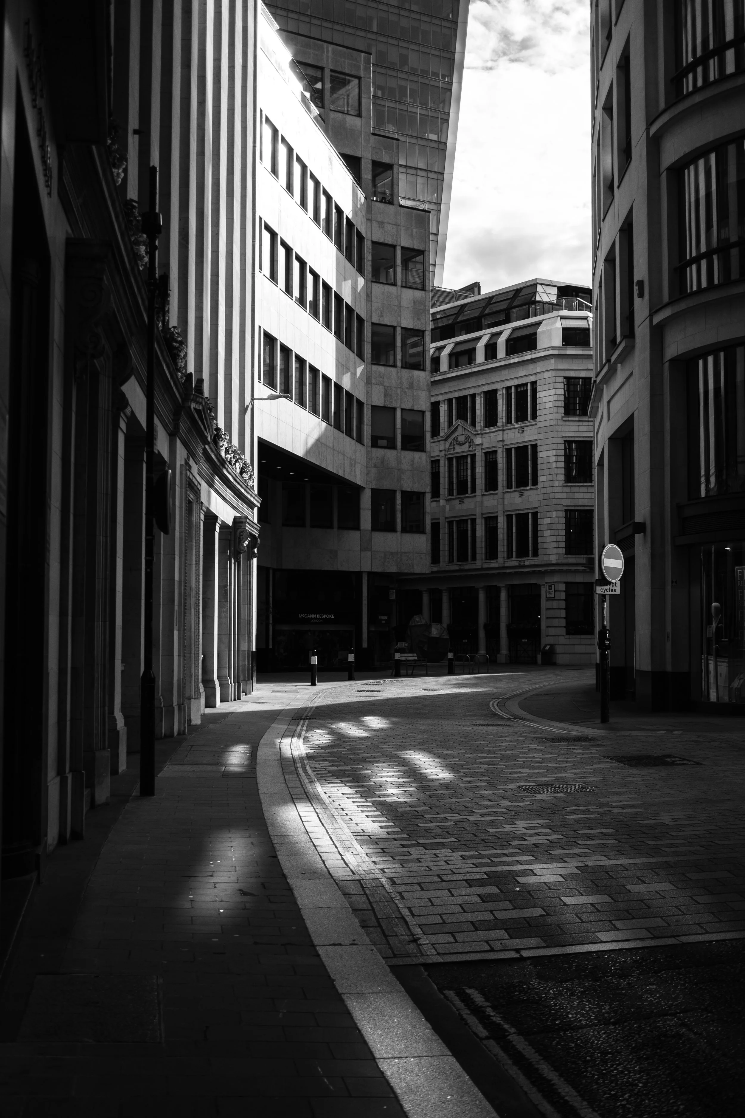 Empty city street with tall modern buildings, some shadows on the cobblestone pavement, in black and white.