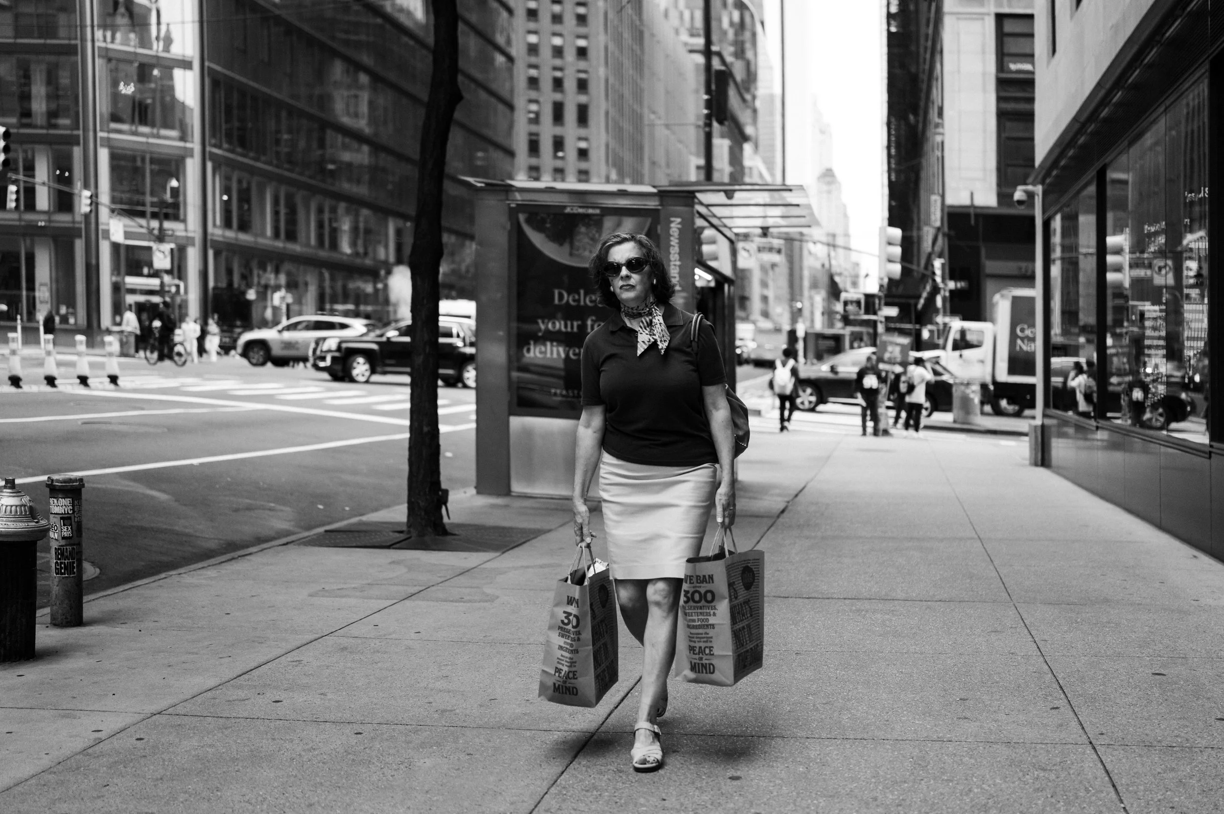 A woman with sunglasses and a scarf around her neck walking on a city sidewalk carrying shopping bags, with tall buildings and other pedestrians in the background.