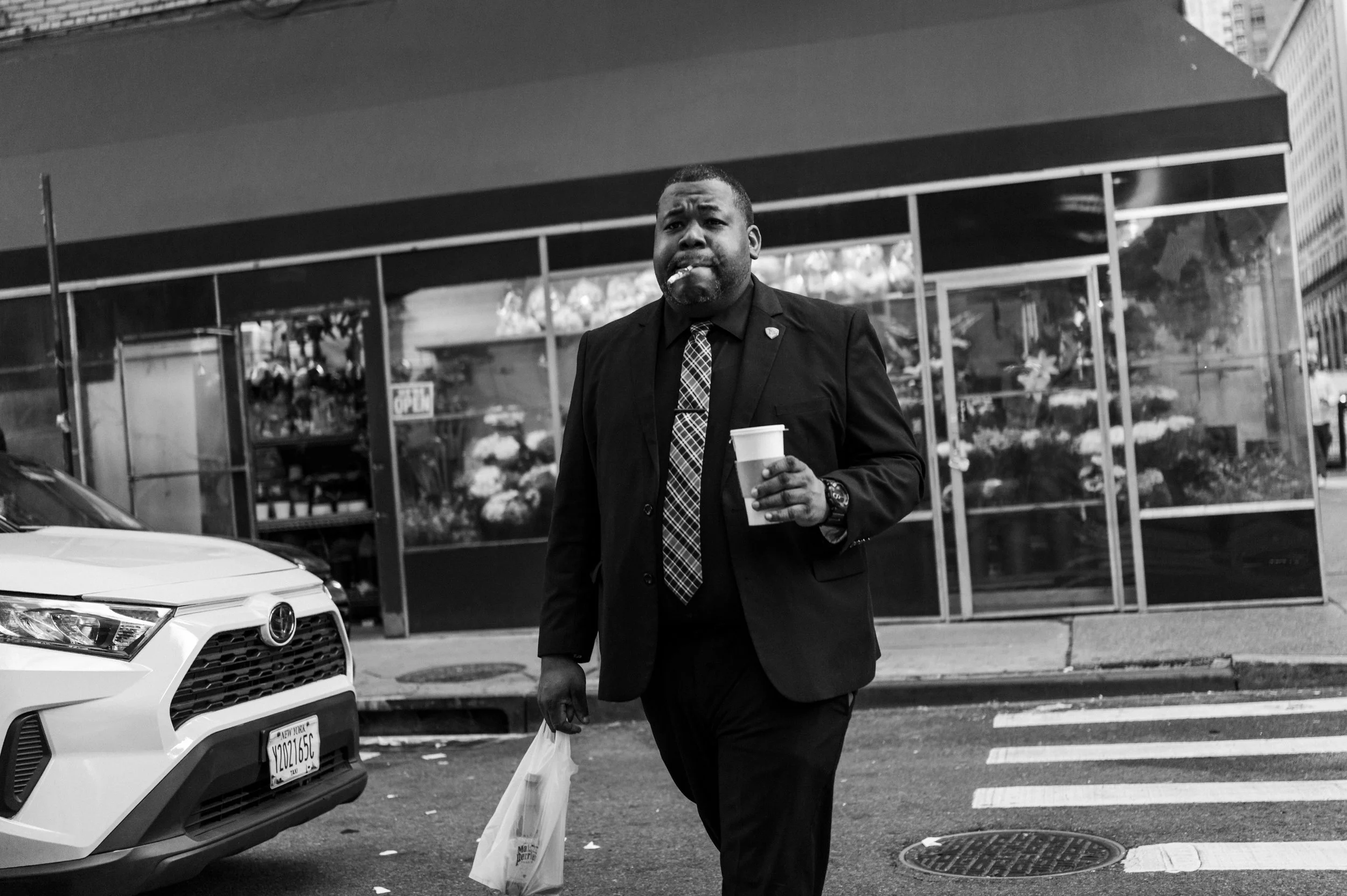 A man in a black suit and patterned tie walking across a street, holding a coffee cup and a plastic bag, with a floral shop and cars in the background.