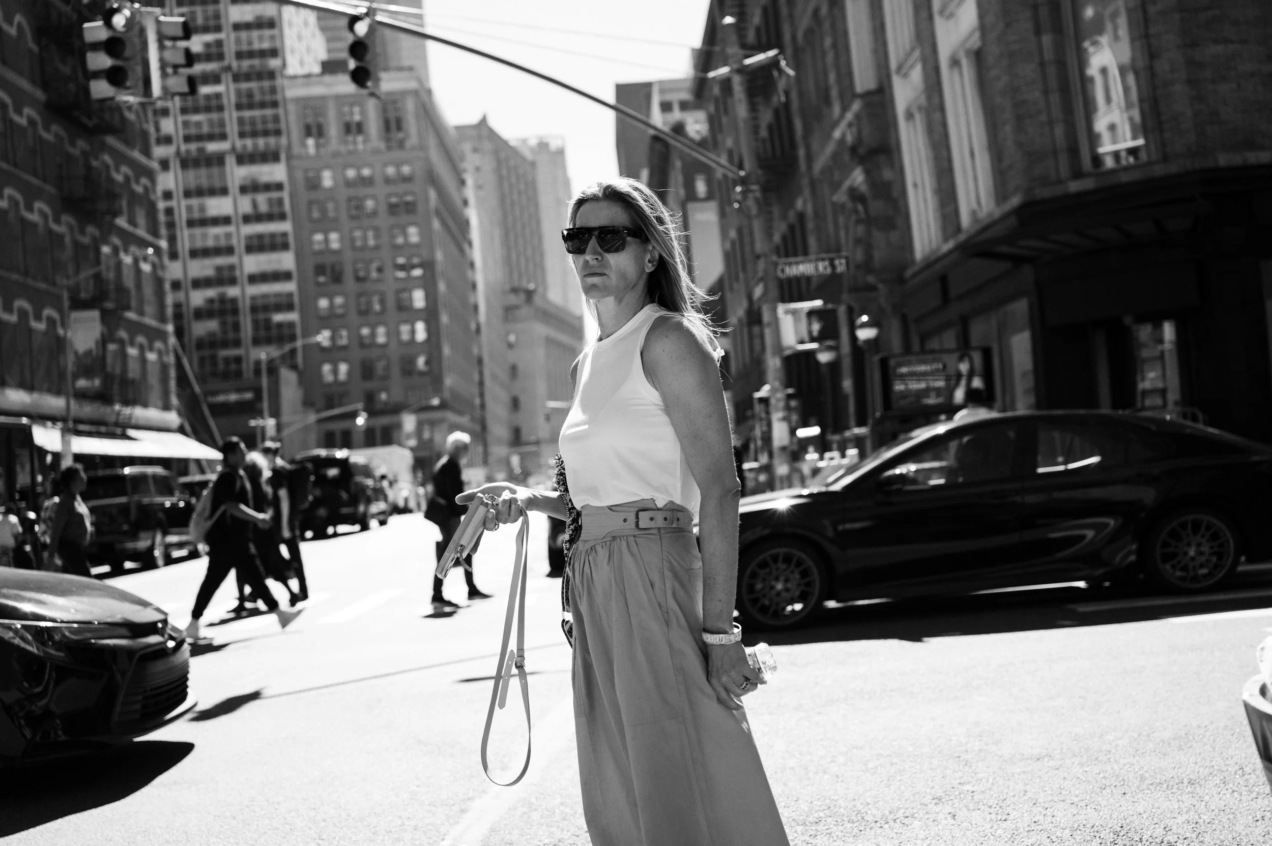 A woman wearing sunglasses and a sleeveless white top standing on a city street corner, holding a leash and possibly a water bottle, with cars and pedestrians in the background.