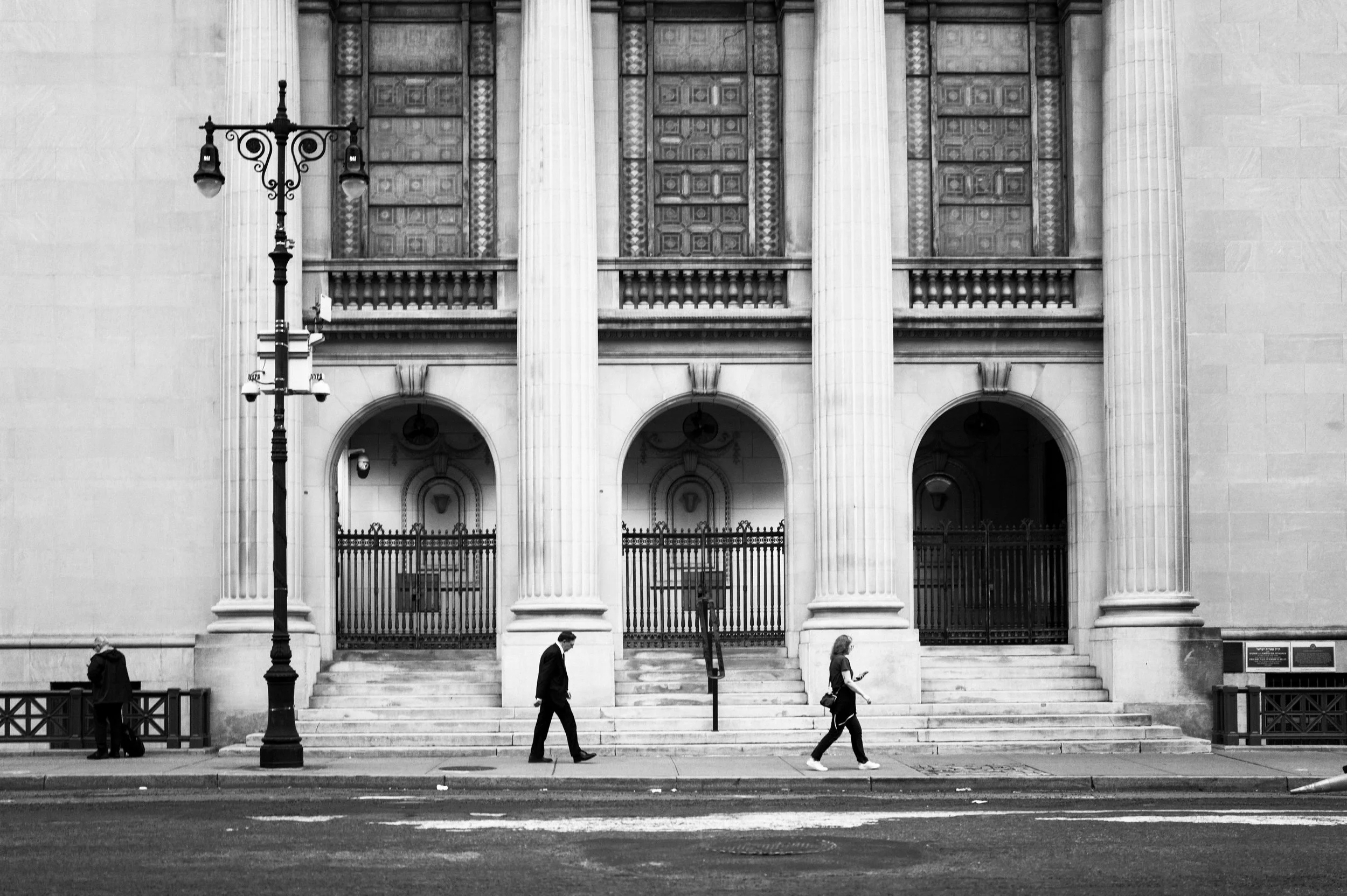 Black and white photo of a church or government building with large columns, stairs, and ornate doors, a lamppost in the foreground, and three people walking on the sidewalk.