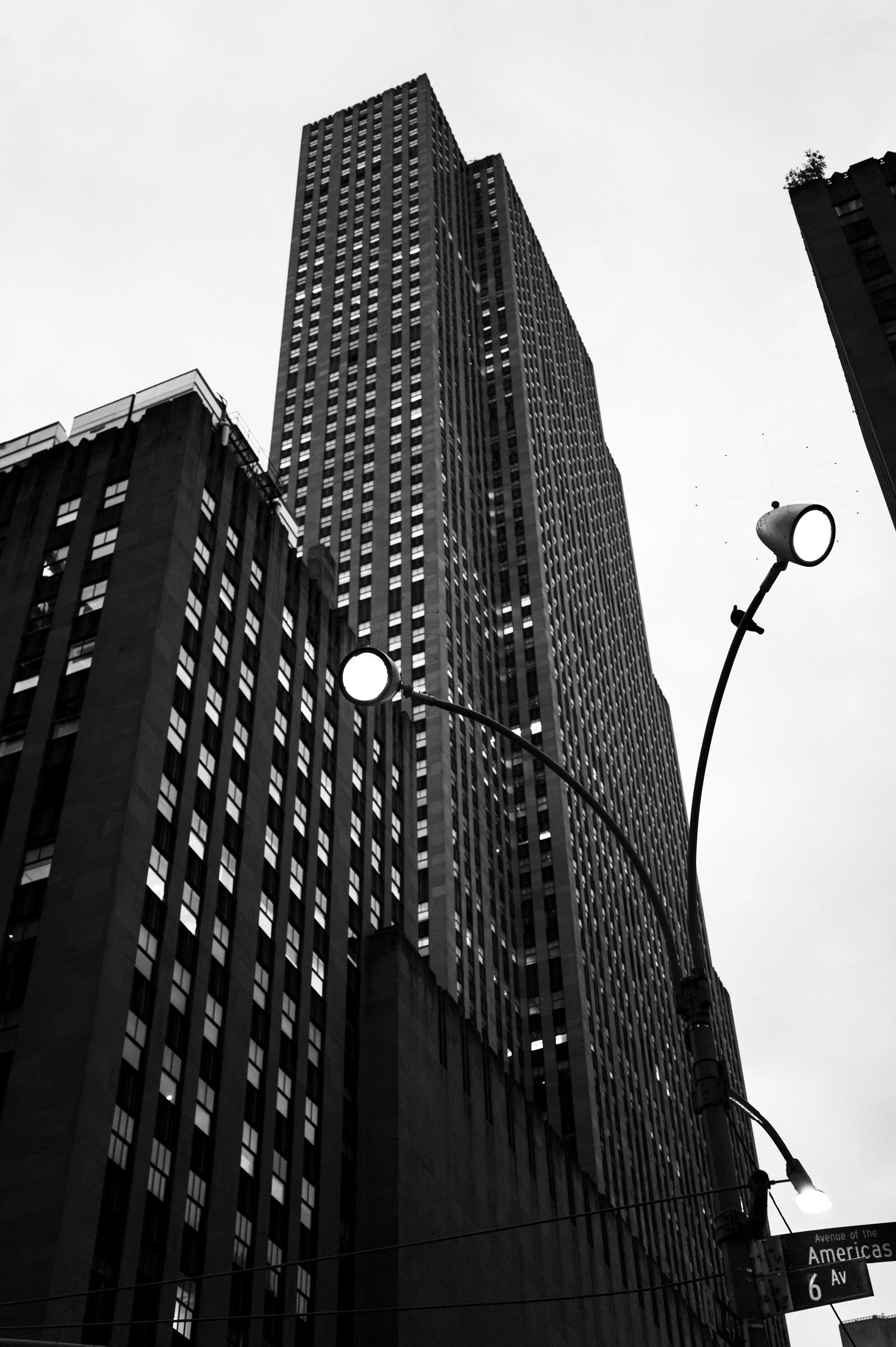 Black and white photo of tall skyscrapers in an urban cityscape, with street lamps and a street sign reading 'Avenue of the Americas, 6 AV' in the foreground.