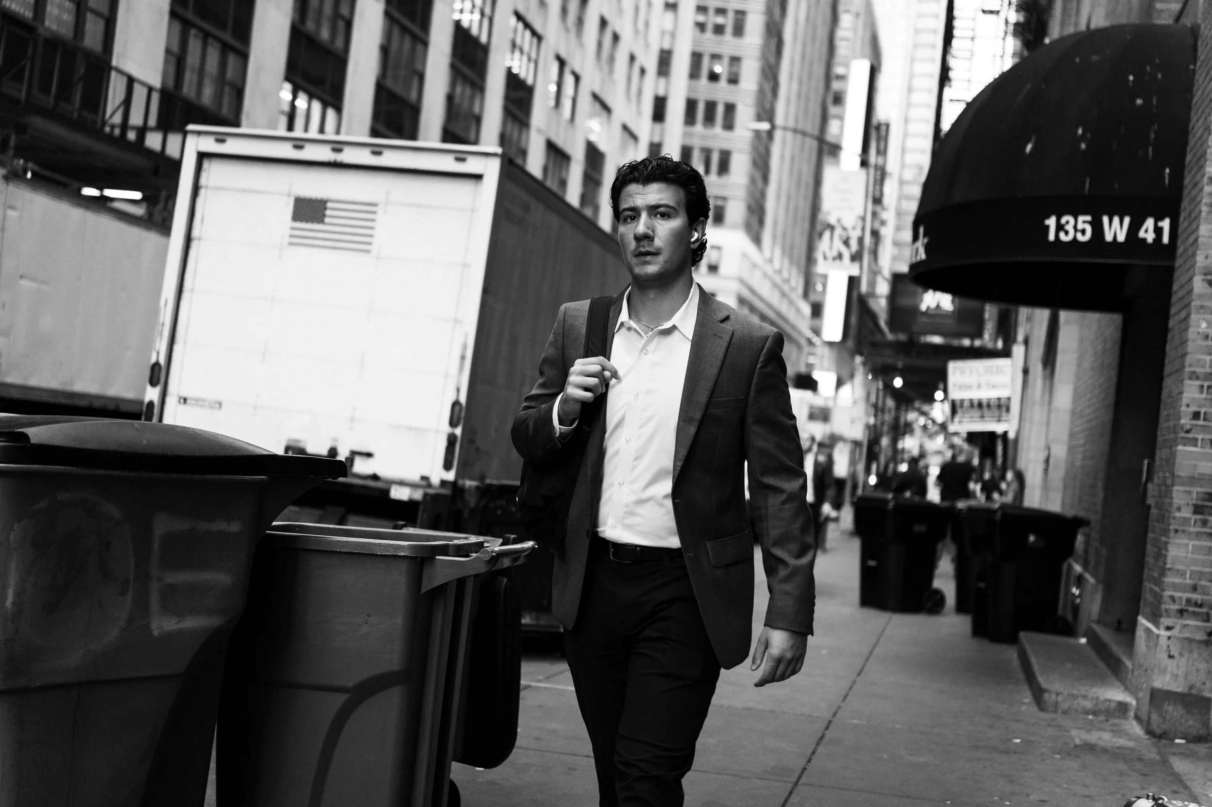 A young man in a business suit walking down a city sidewalk carrying a shoulder bag, with buildings and a large delivery truck in the background.