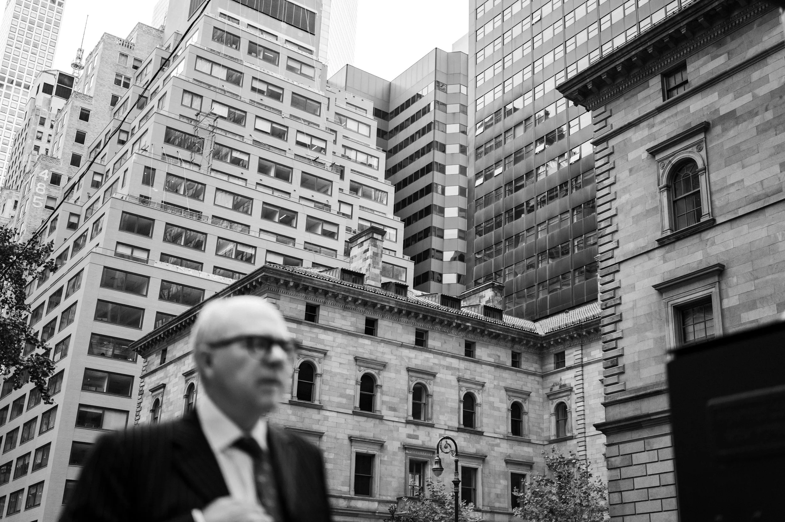 A blurred man in a suit and glasses standing in front of tall modern and historic buildings in an urban cityscape.