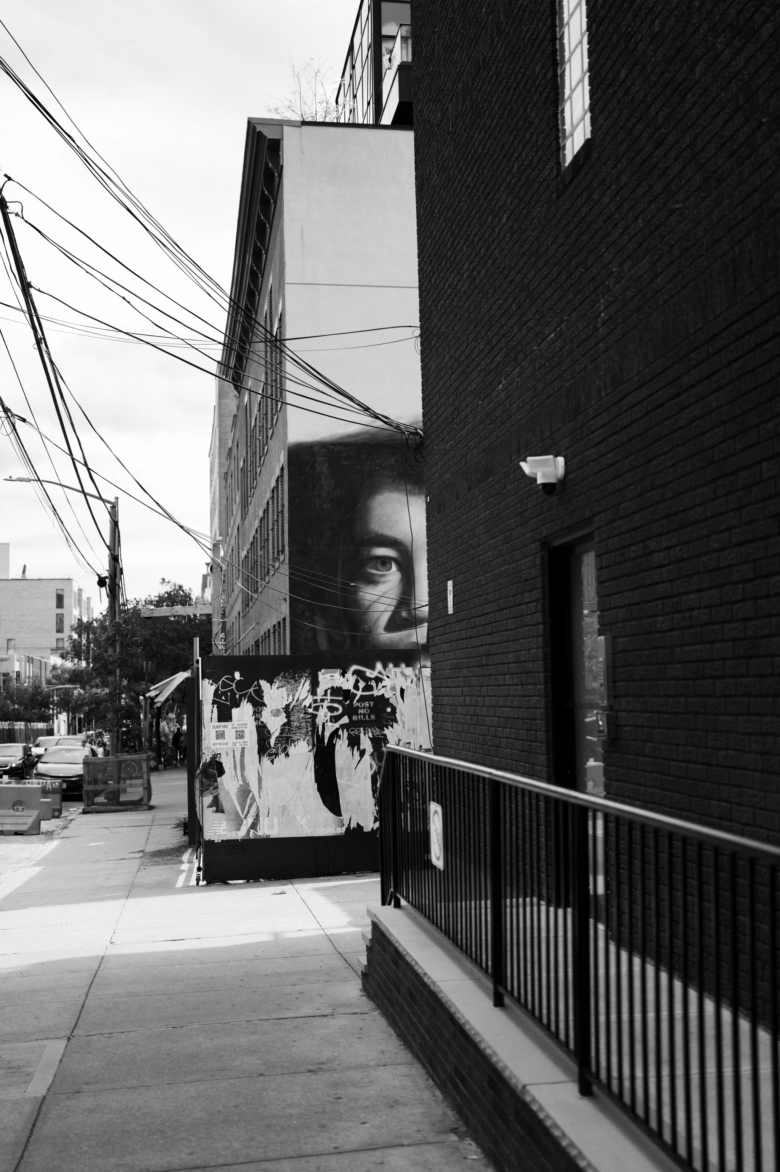 A black and white photo of an urban street scene. A large mural of a woman's face with only one eye visible is painted on the side of a building. There are power lines crossing the sky and parked cars on the street.
