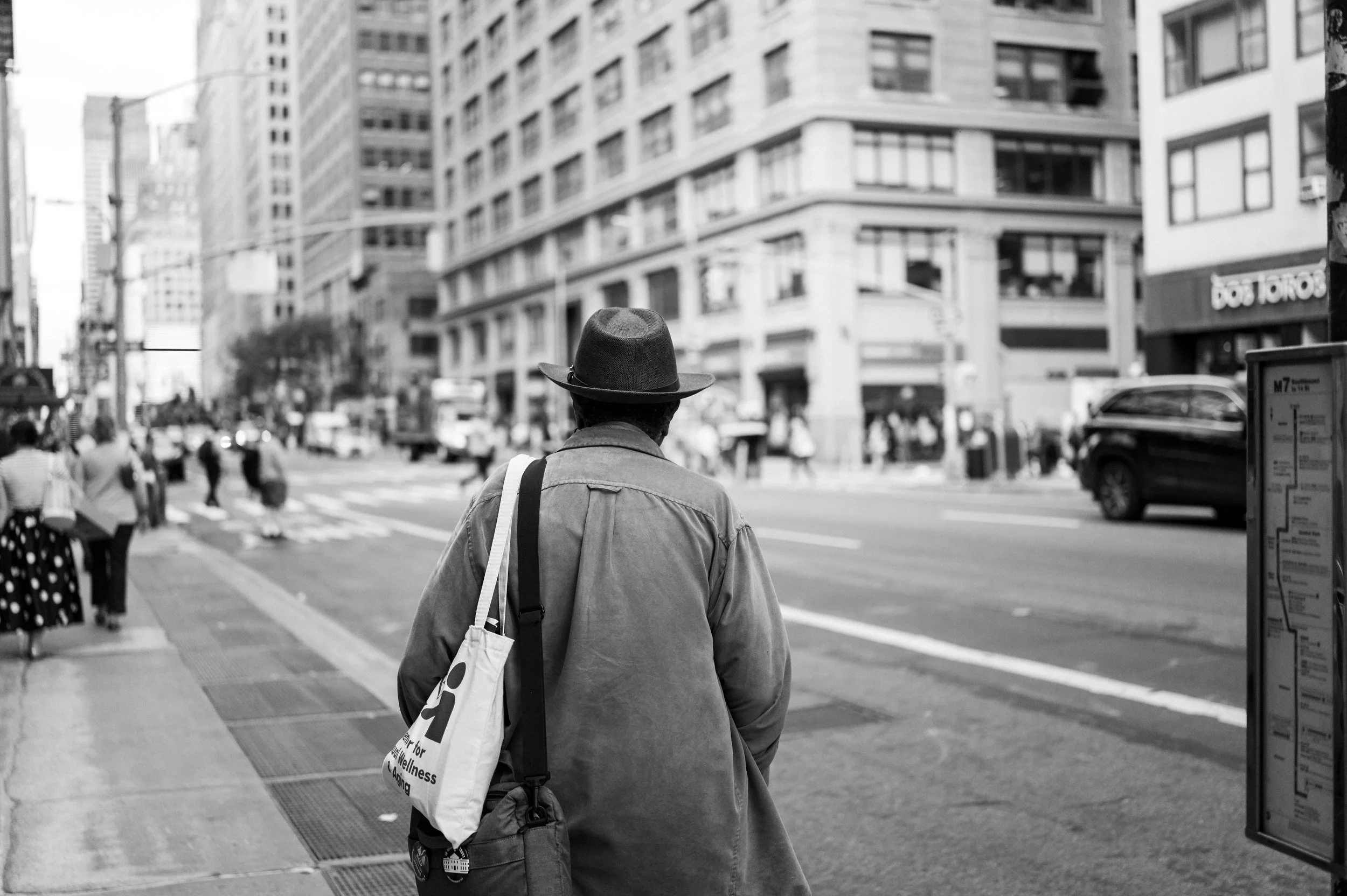 A person wearing a hat and a jacket, carrying a tote bag, walking on a busy city street in black and white.