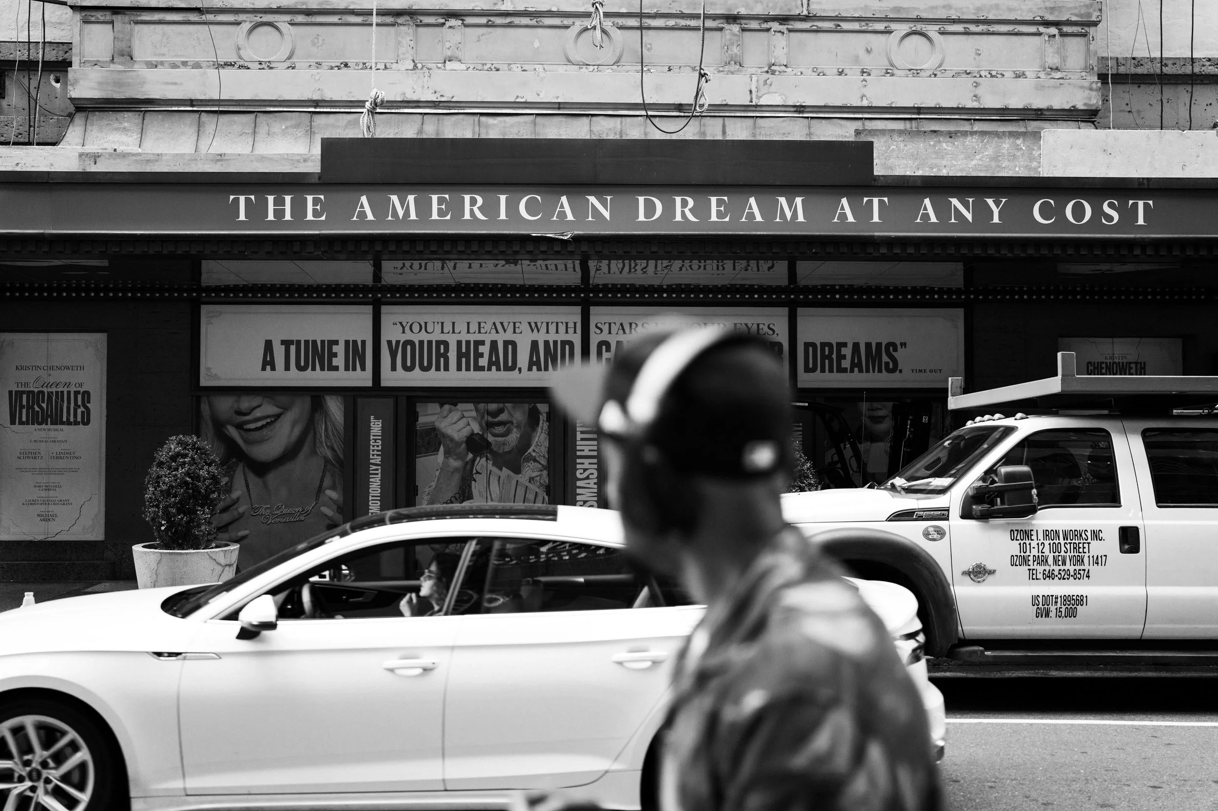 Black and white street scene with a person in a hat and hoodie walking past a line of cars parked in front of a theater. The theater's marquee reads "The American Dream at Any Cost" and features a quote about leaving with dreams, along with posters a