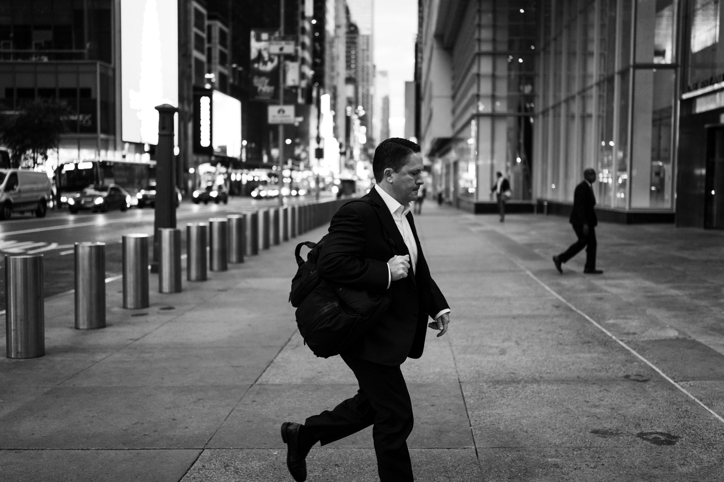 Man in suit with backpack walking across city sidewalk in black and white