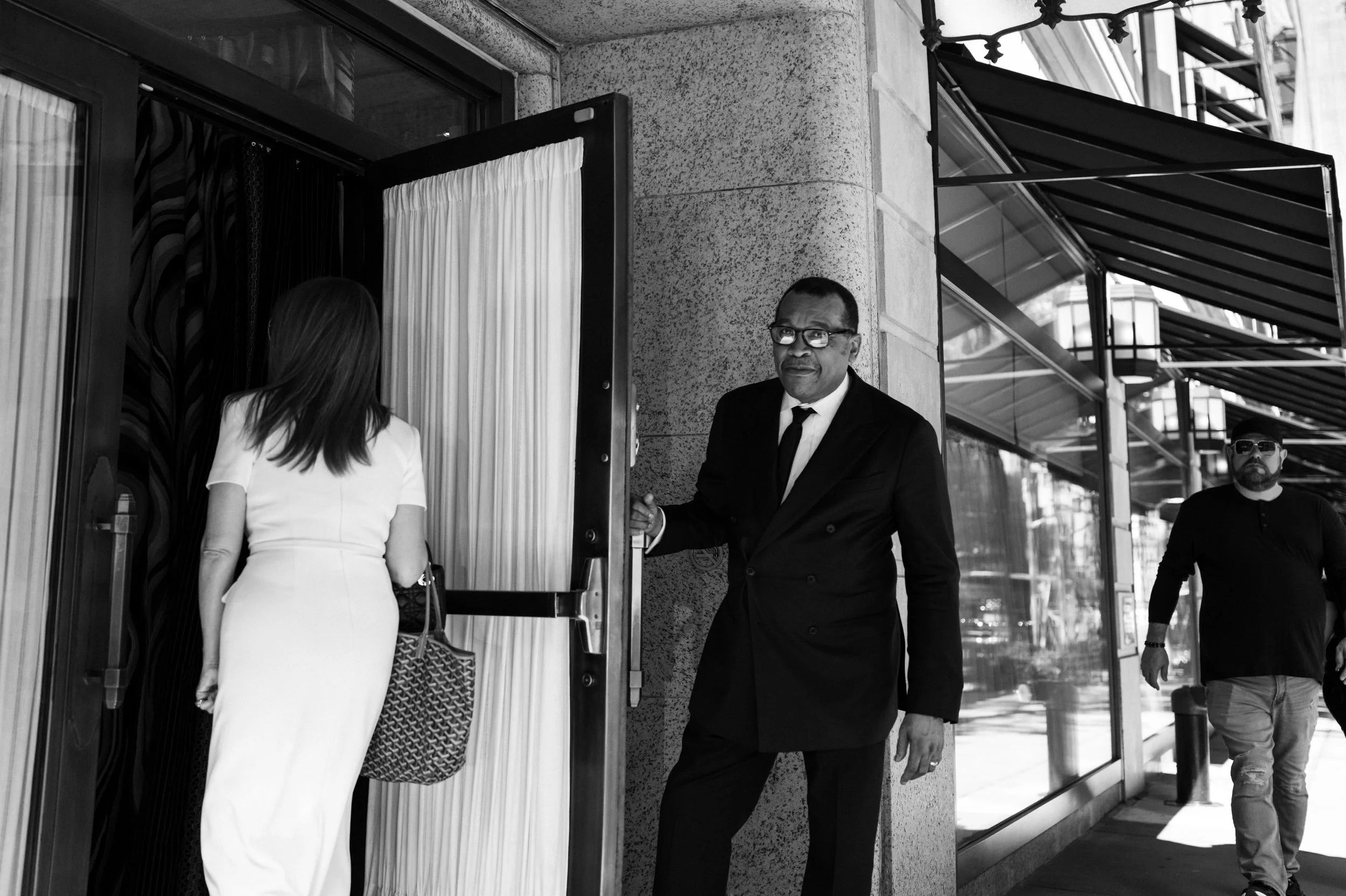 A man in a black suit and glasses standing outside a building, holding the door open for a woman entering. The woman is wearing a white dress and carrying a patterned bag. Another man is walking by in the background.