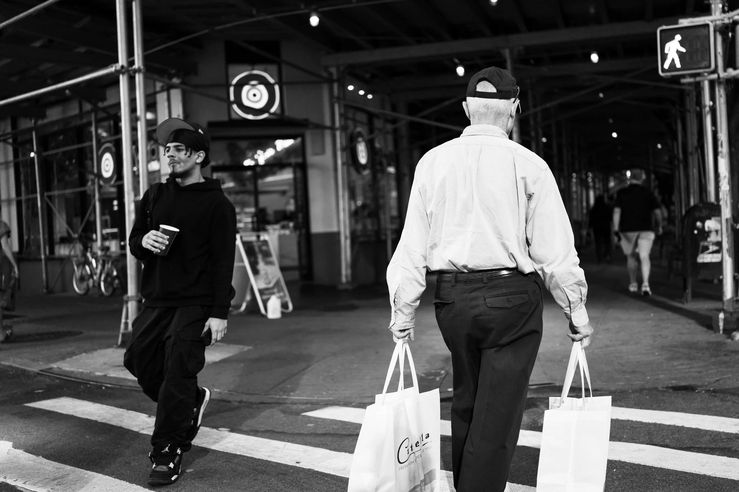 An older man and a younger man crossing a city street at night. The older man is walking away from the camera, holding shopping bags, while the younger man is walking toward the camera, holding a cup. City lights and pedestrians are visible in the ba