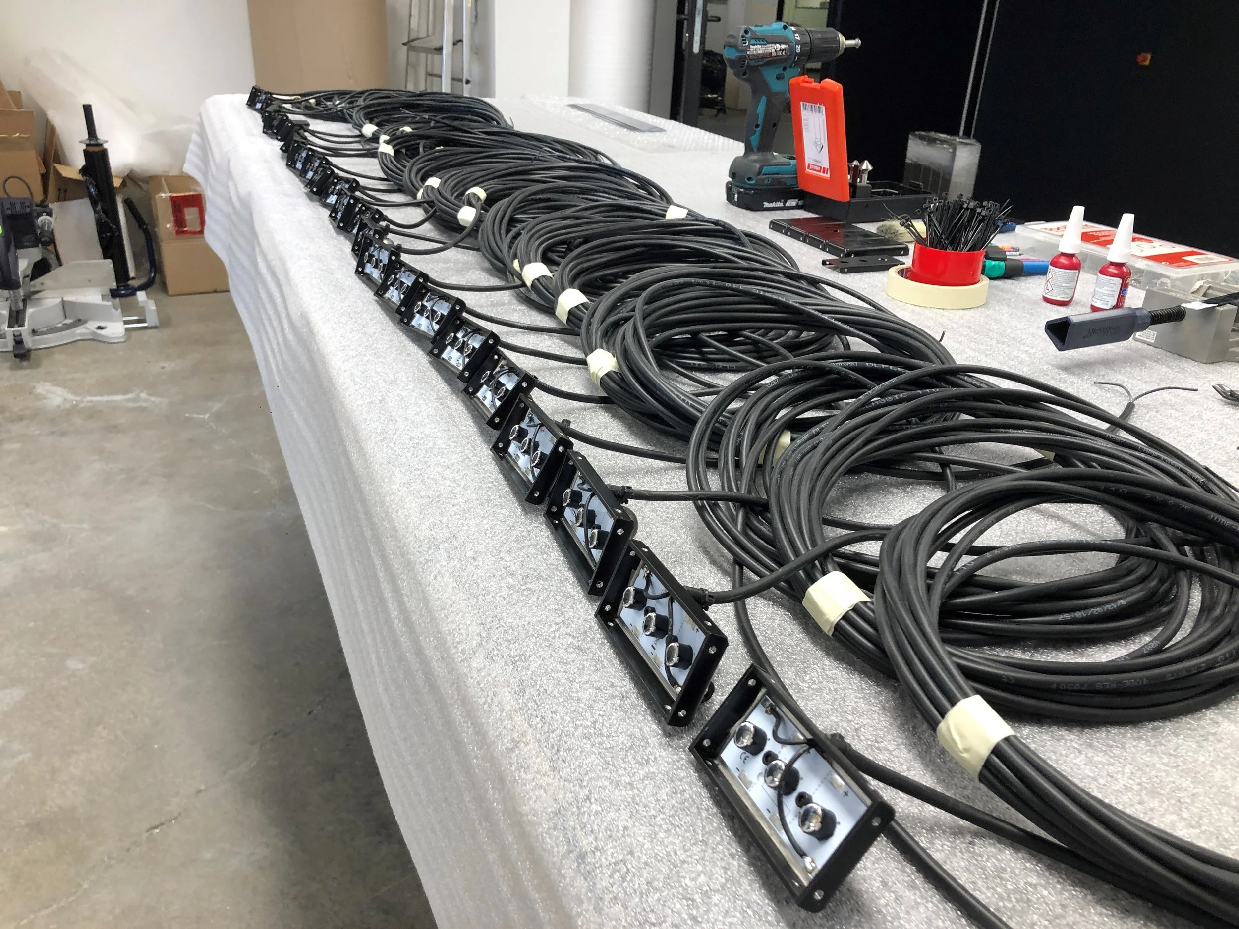 Serienproduktion, Arbeitsstraße, viele schwarze Kabel liegen auf einem Tisch, einige sind mit Schaltern versehen. Es sind Werkzeuge und Kleinteile auf dem Tisch, GODEV Werkstatt.