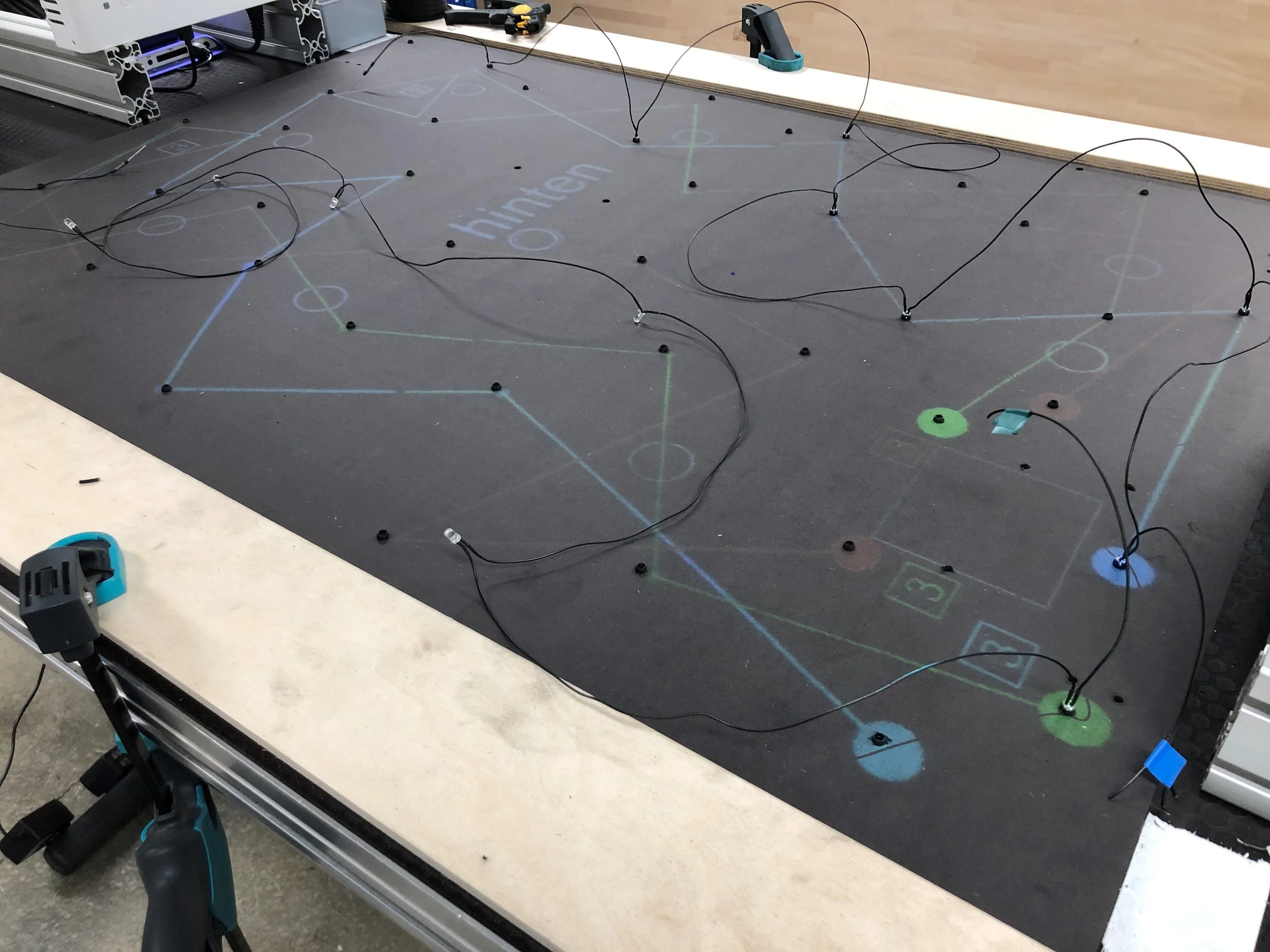 Auf einem schwarzen Spielfeld sind mehrere Lichter und Kabel angebracht, die für  Robotik- verwendet werden, mit einer Kamera und weiteren technischen Geräten im Hintergrund.