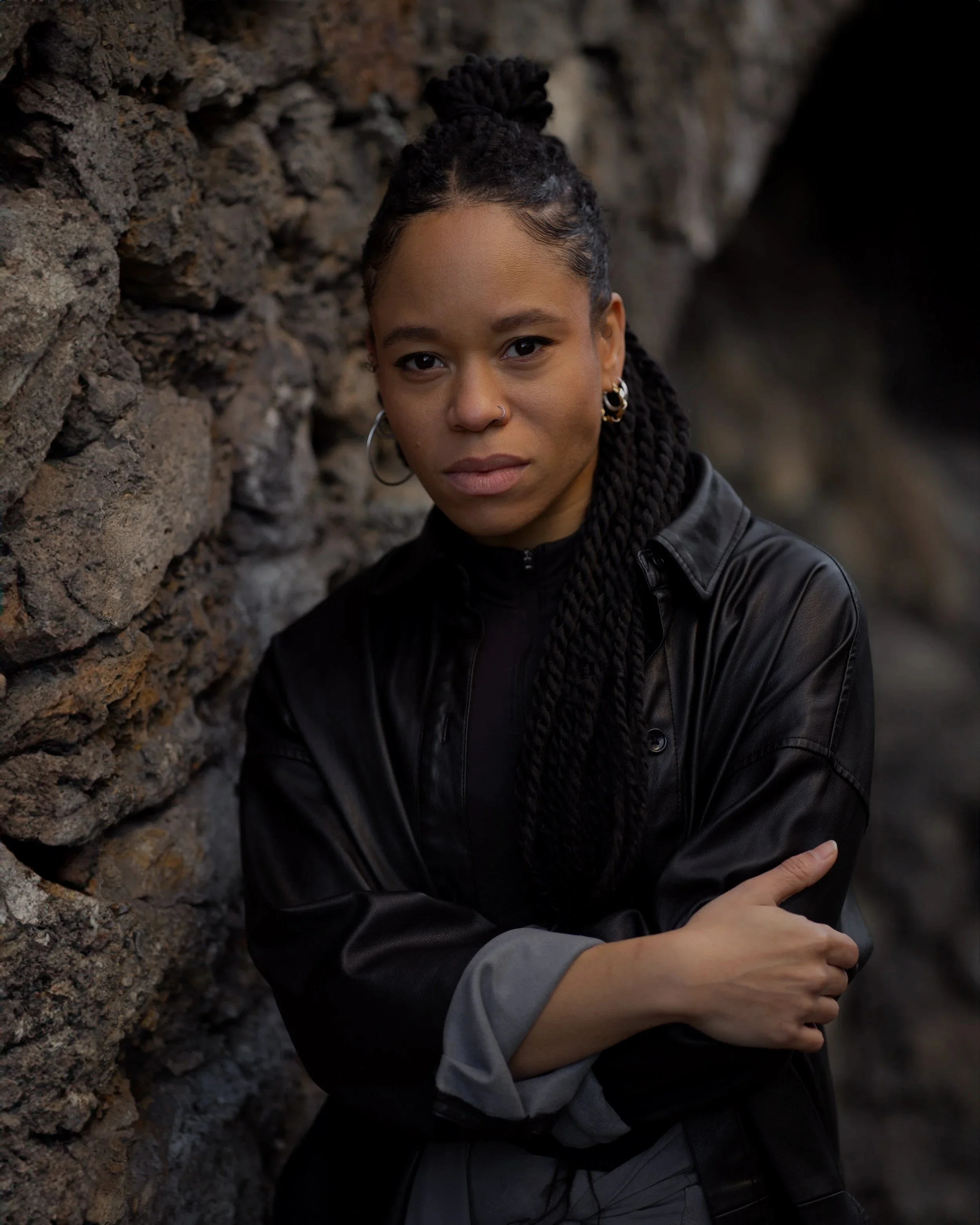 A young woman with braided hair, hoop earrings, and a nose piercing posing against a stone wall, wearing a black leather jacket.