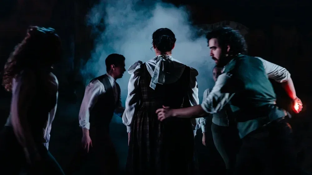 A group of people, mostly men, gathered in a circle in a dark setting with fog and dramatic lighting, engaging in a discussion or activity.