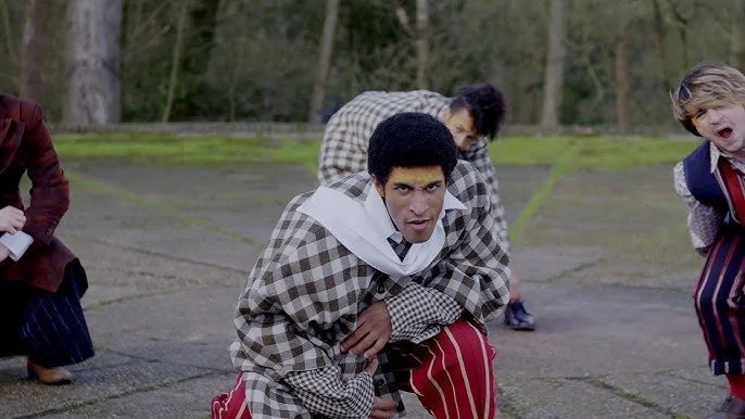 A group of young boys dressed in colorful vintage clothing, crouching outdoors on a paved area surrounded by trees, with serious or intense expressions.