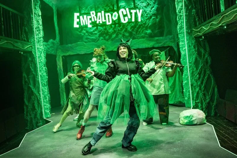 Group of performers dressed in whimsical costumes on stage with a green forest backdrop, holding toy instruments, and a sign reading 'Emerald City' hanging above them.