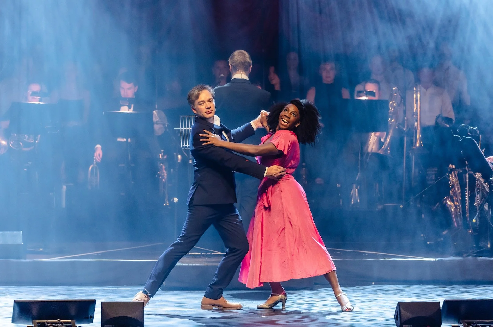 A man and woman are dancing together on stage, with the woman smiling broadly, wearing a pink dress and heels, while the man is in a dark suit. A band with musicians and song instruments is visible in the background, illuminated by stage lights and fog.