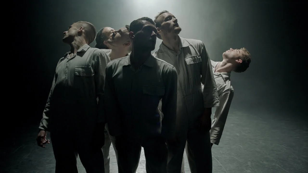 Six men in work uniforms stand in a dark, moody setting, looking upwards with varied expressions.