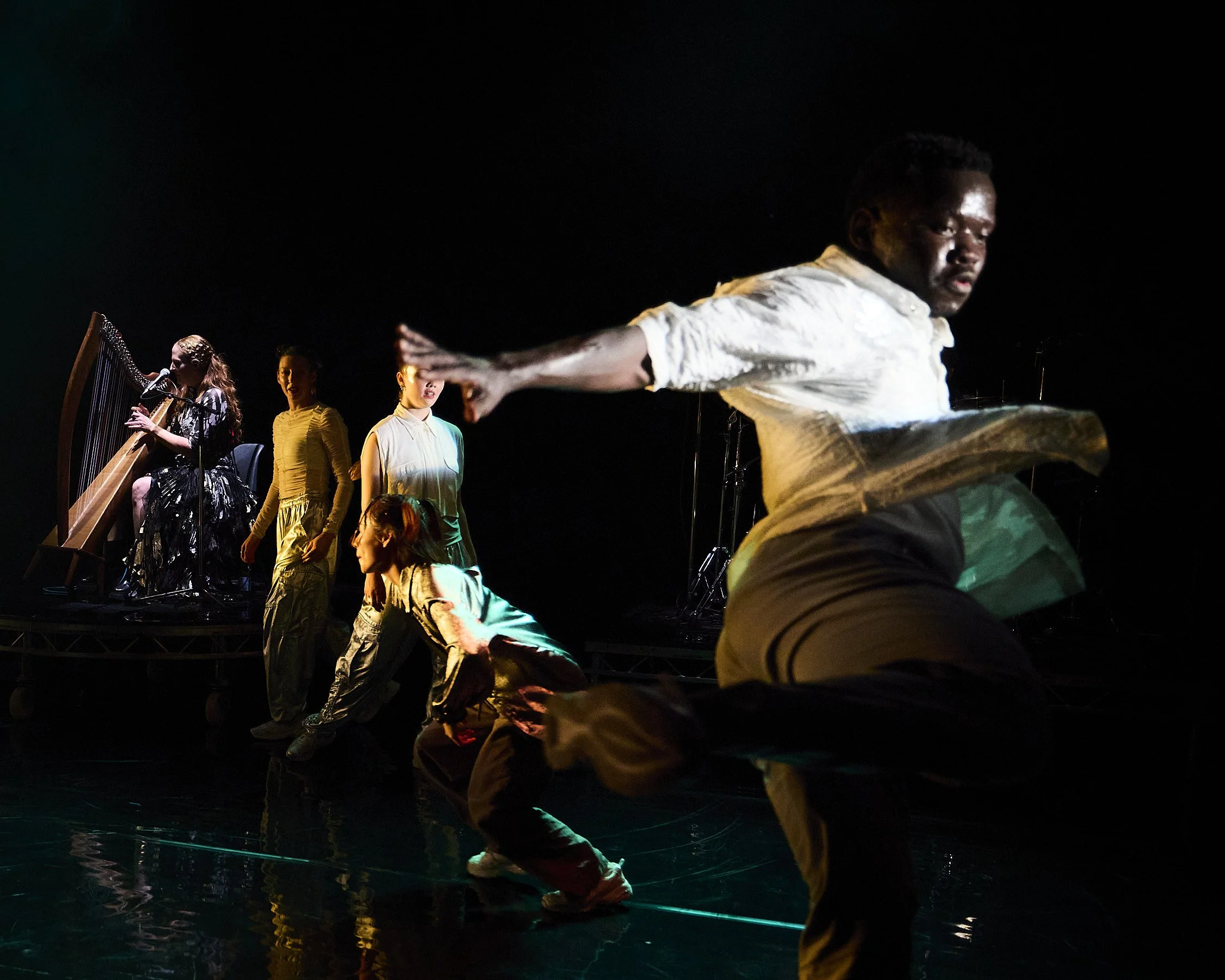 A group of five performers on stage, with one playing a harp, two standing, one crouching, and one in a dynamic pose, all illuminated against a dark background.
