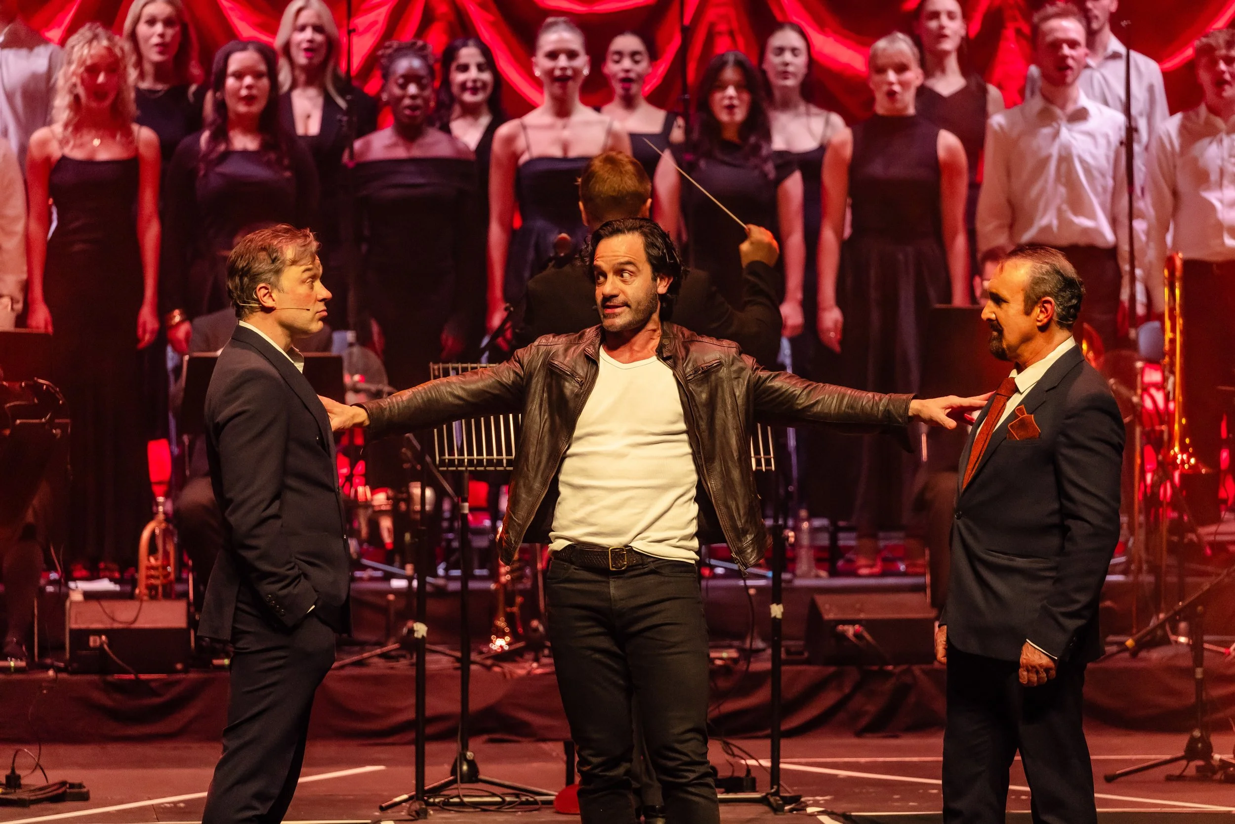 Stage scene with three men, one in a leather jacket and two in suits, engaging in conversation, surrounded by a choir and orchestra in the background.